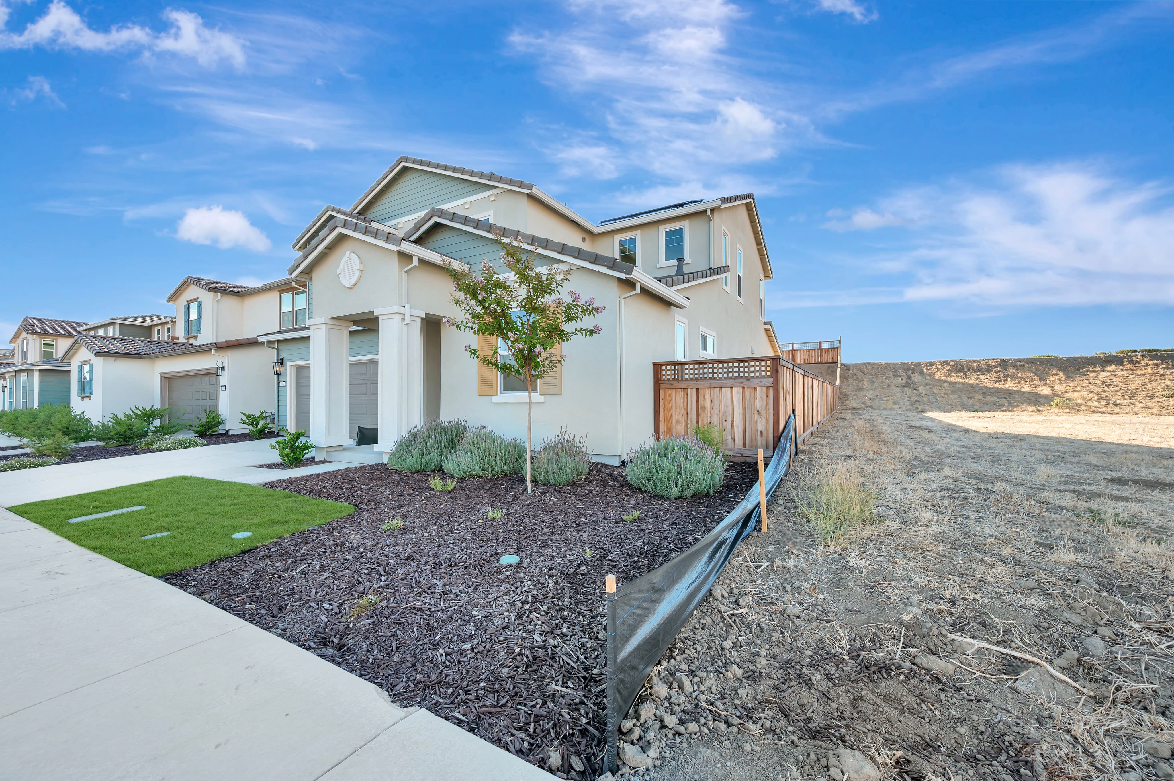A modern beige house with a manicured lawn and landscaping sits in the foreground, while a vast, undeveloped hillside stretches into the background under a bright blue sky.