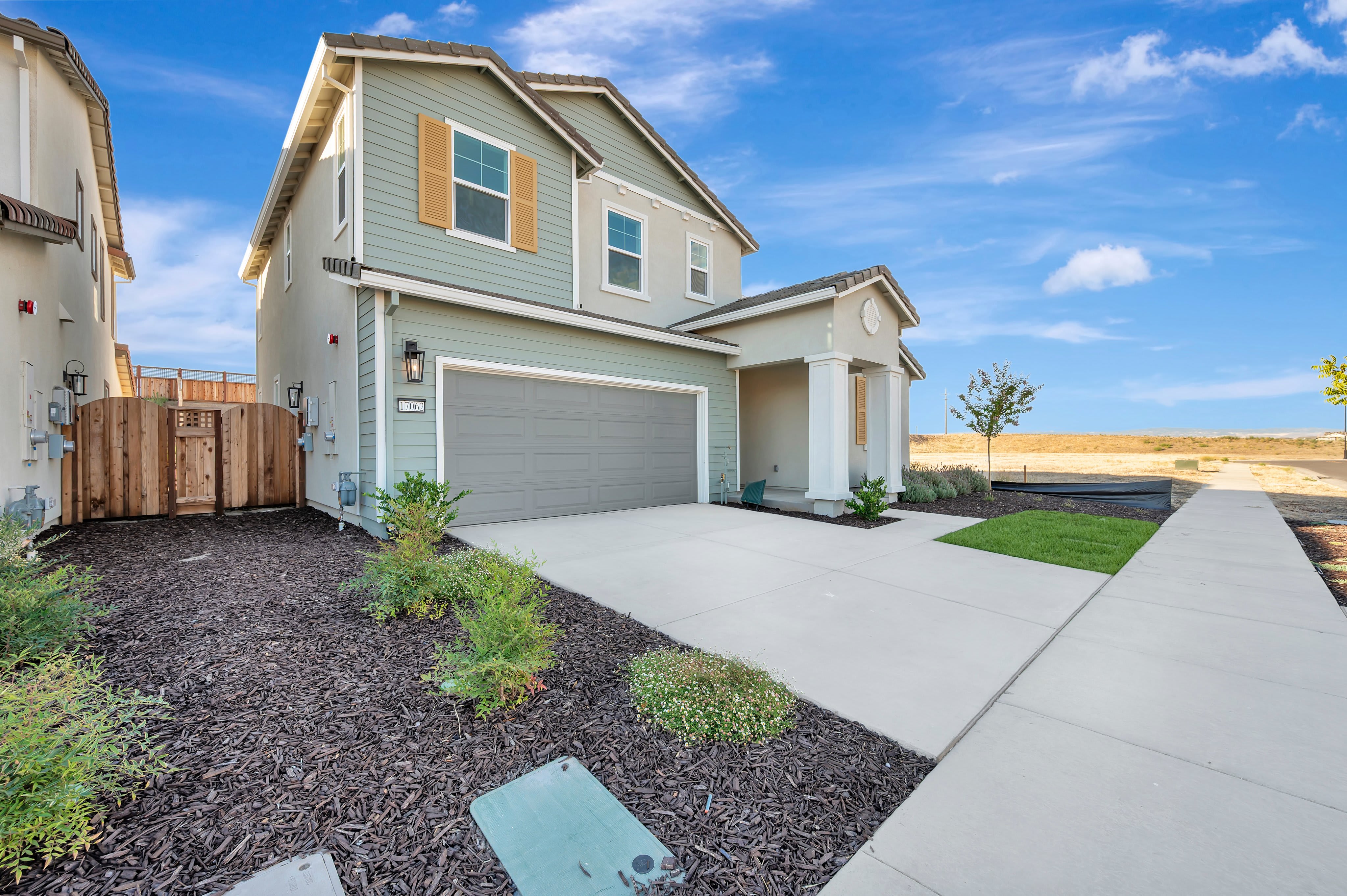 A modern two-story house with a gray garage door and light green siding sits on a concrete driveway, with a sidewalk leading to a vast, undeveloped field under a bright blue sky.