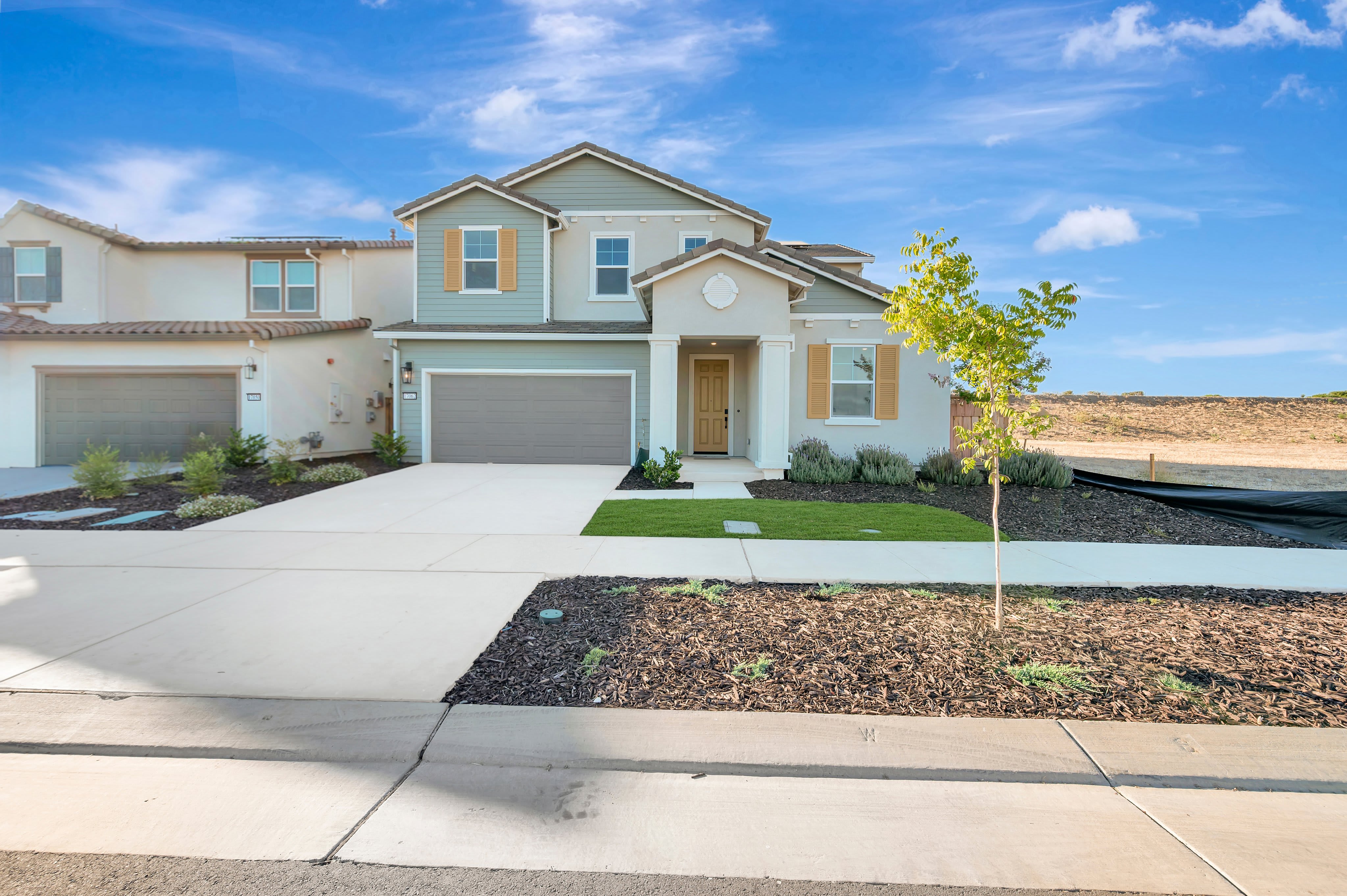A modern townhouse with a manicured lawn and driveway in the foreground is set against a backdrop of a clear blue sky and undeveloped land.