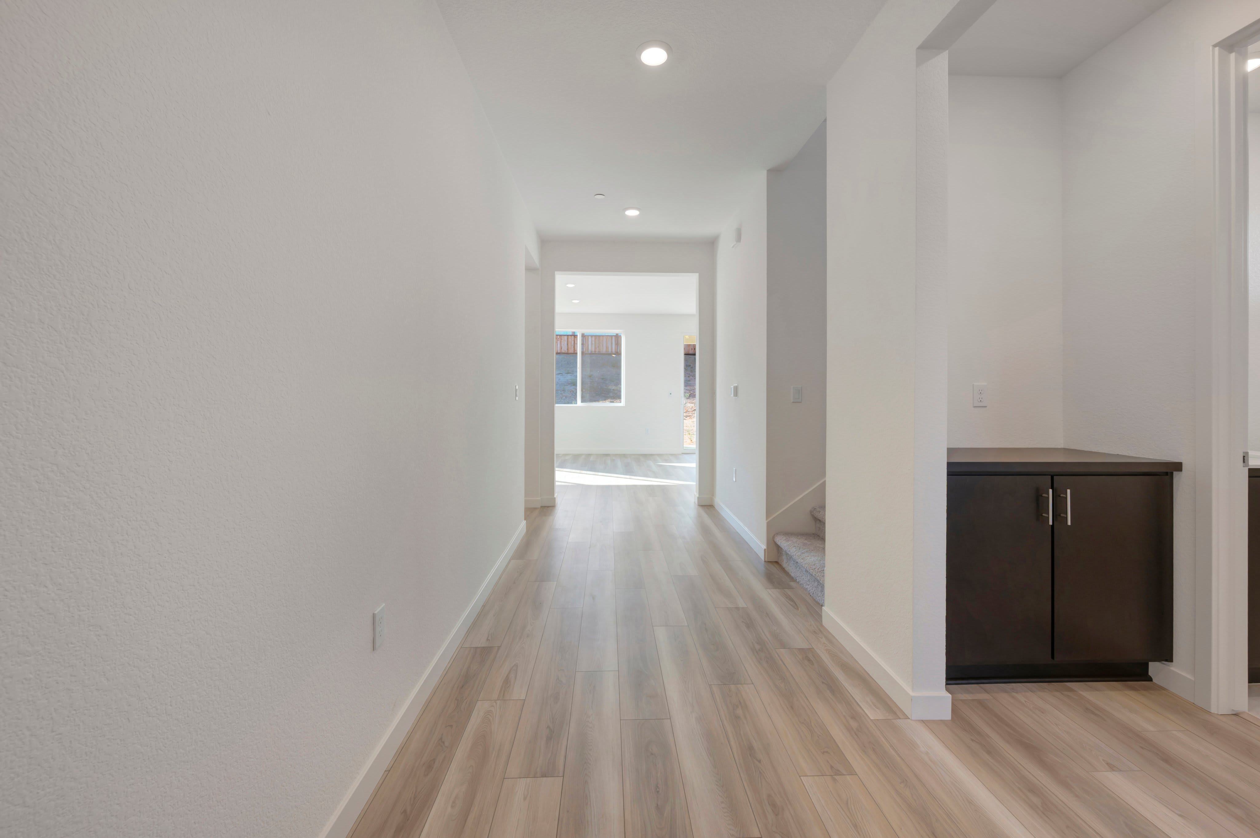 A bright, modern hallway with light wood flooring in the foreground leads to a sunlit room with a window in the background.