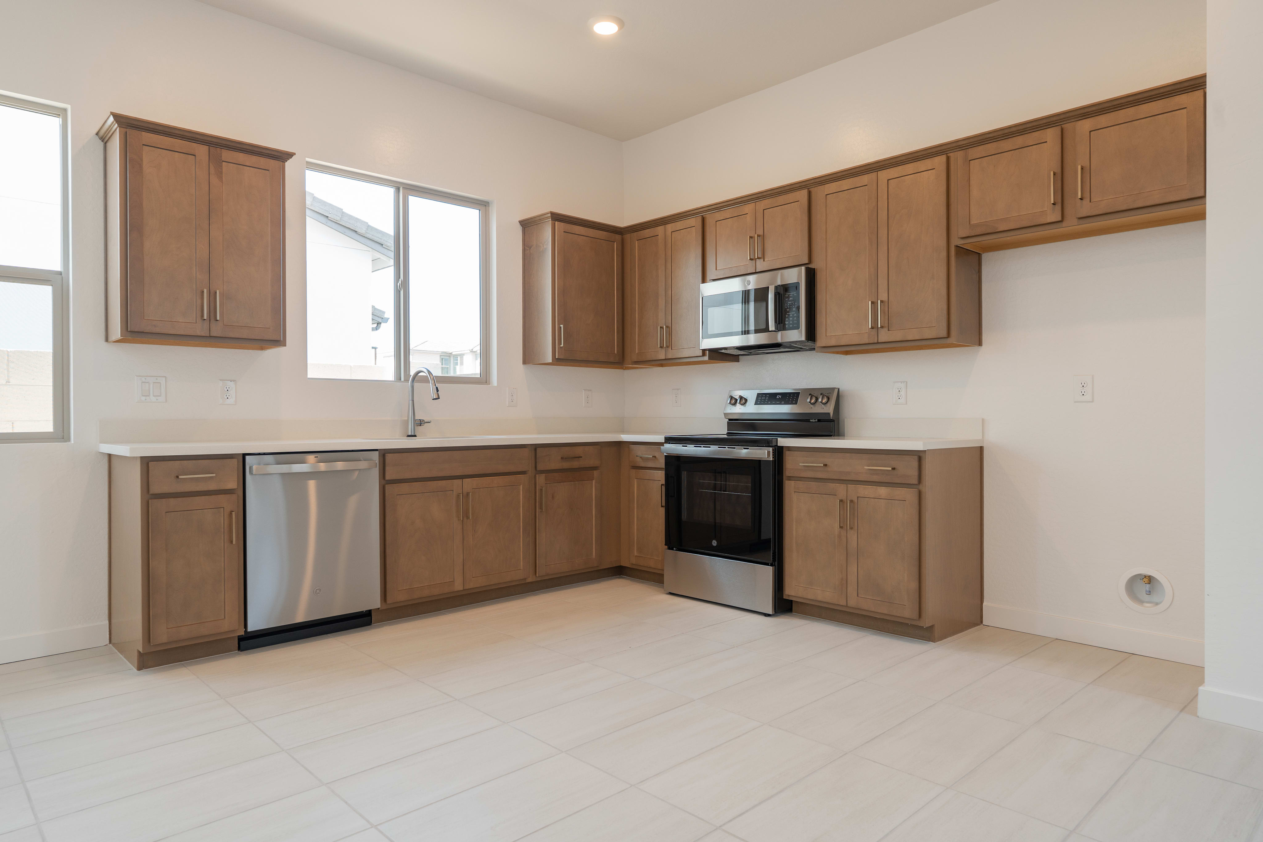 A modern kitchen with a stainless steel dishwasher and oven in the foreground and light brown cabinets and windows in the background.
