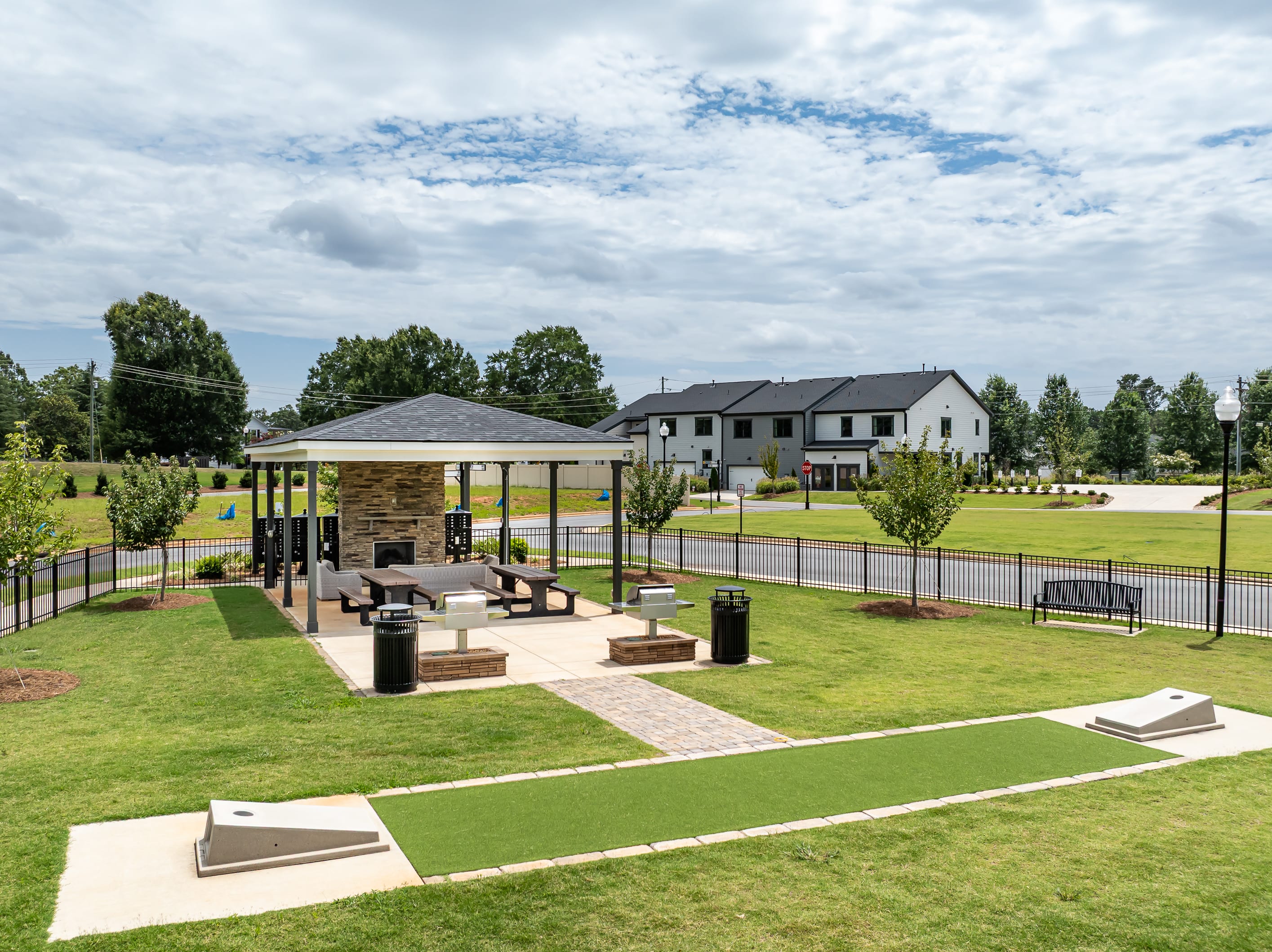 A covered picnic area with grills and tables sits in the foreground, while modern townhouses and a treed landscape form the background under a cloudy sky.