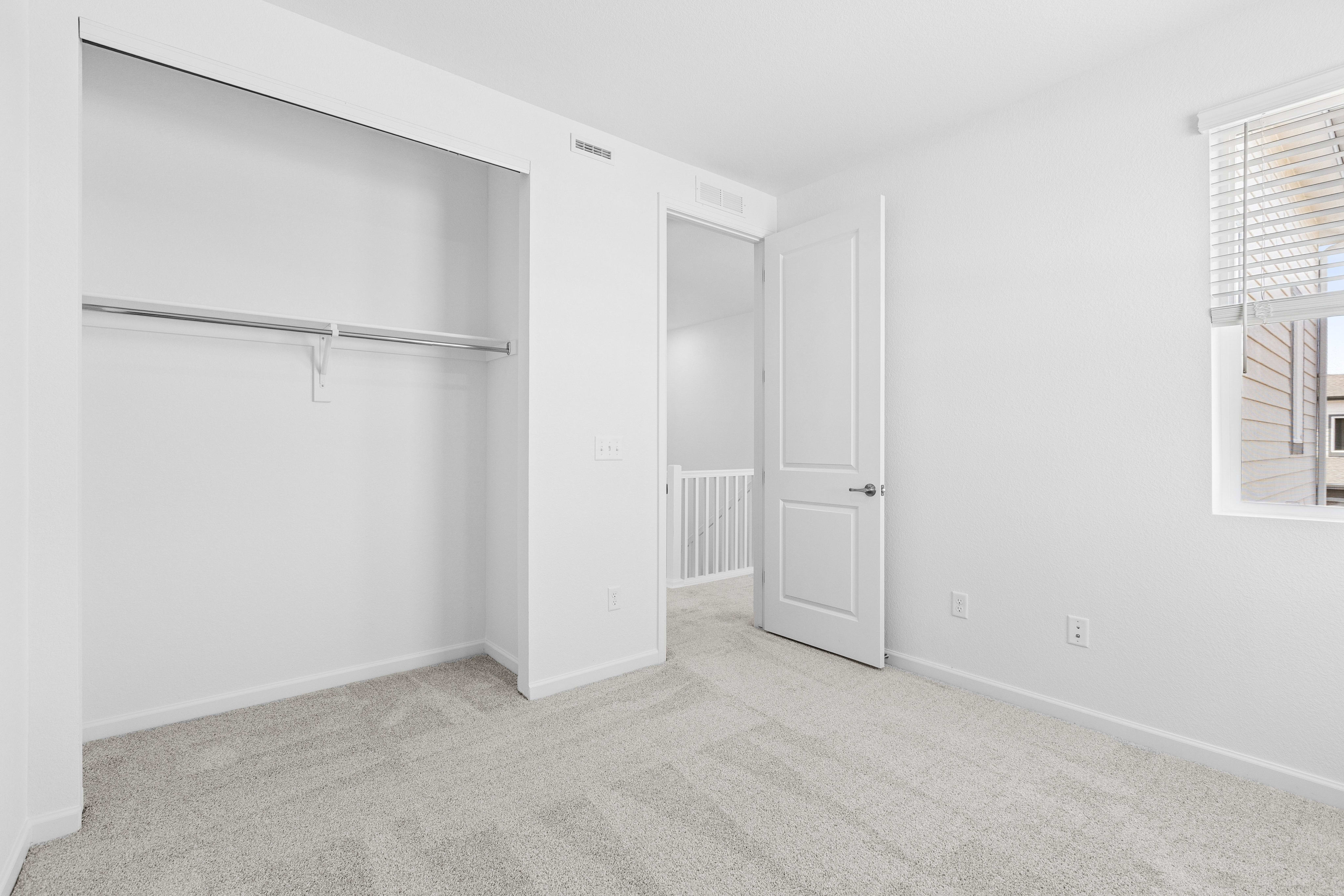 A bright, empty bedroom with a closet in the foreground and a hallway with stairs visible through an open door in the background.