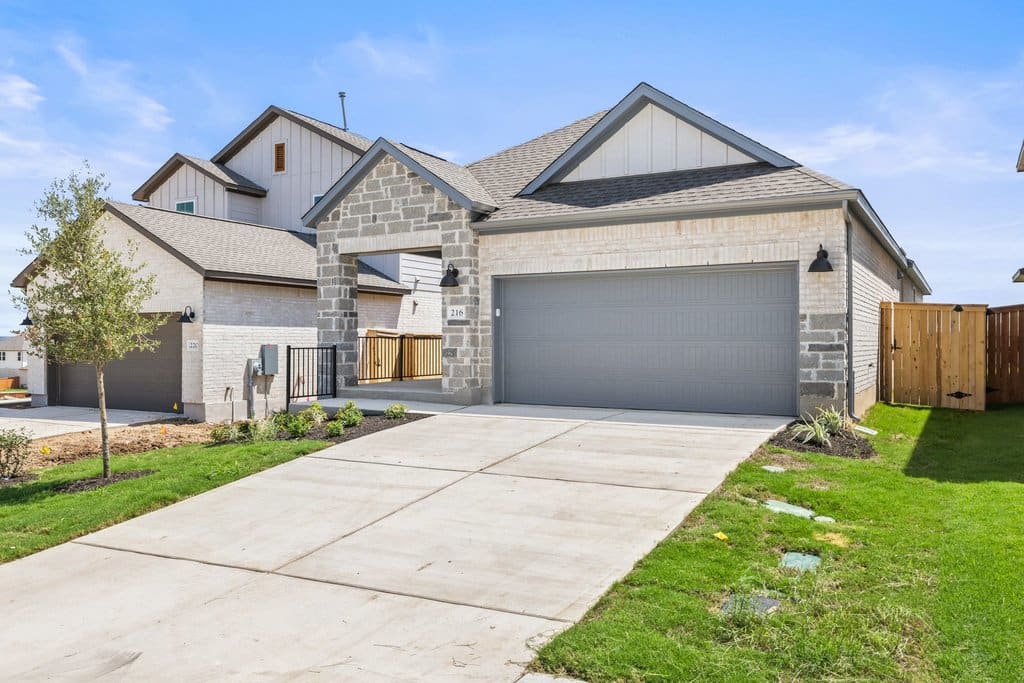 A modern house with a gray garage door and stone accents sits on a concrete driveway, with a bright blue sky and green lawn in the background.
