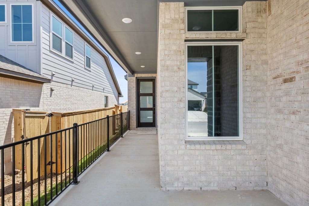 A concrete patio with a black metal railing runs alongside a light brick house with a glass-paneled door, while neighboring houses and a wooden fence are visible in the background.
