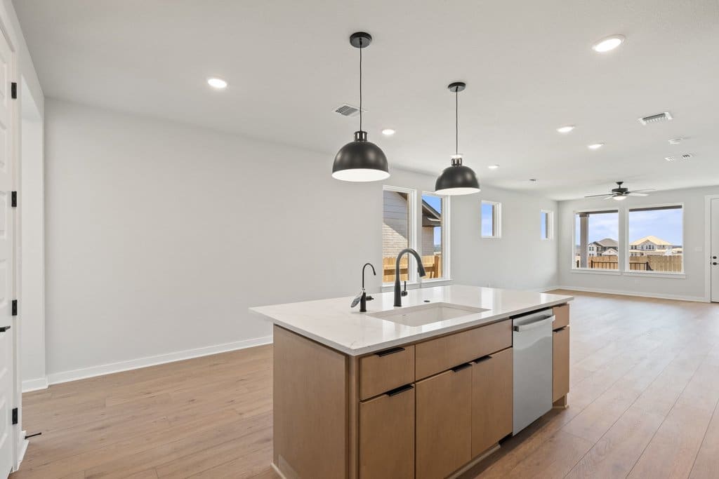 A modern kitchen island with pendant lights in the foreground is set against a bright, open living space with large windows in the background.