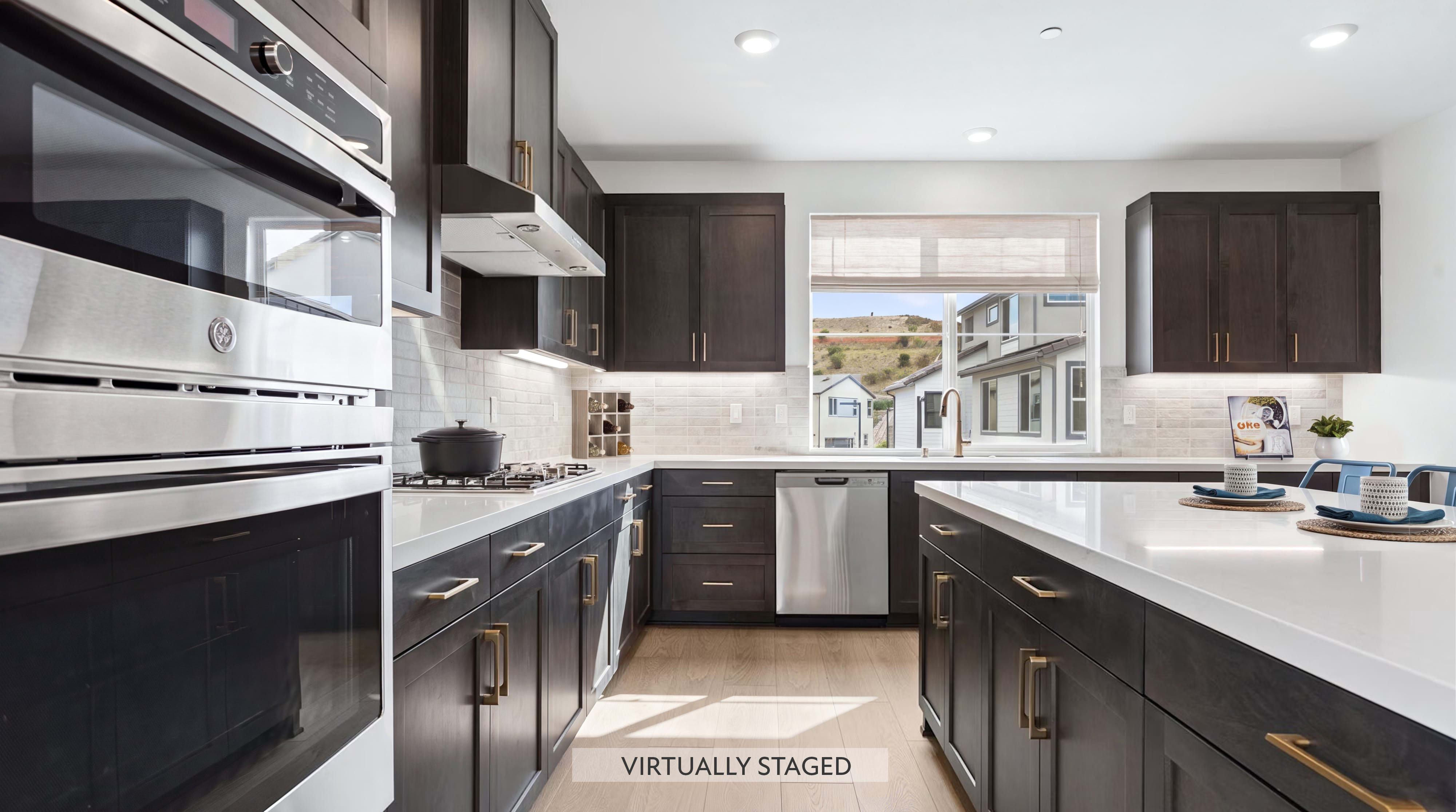 A modern, dark-wood kitchen with stainless steel appliances and a white island is brightly lit, with a view of houses and hills through the window in the background.