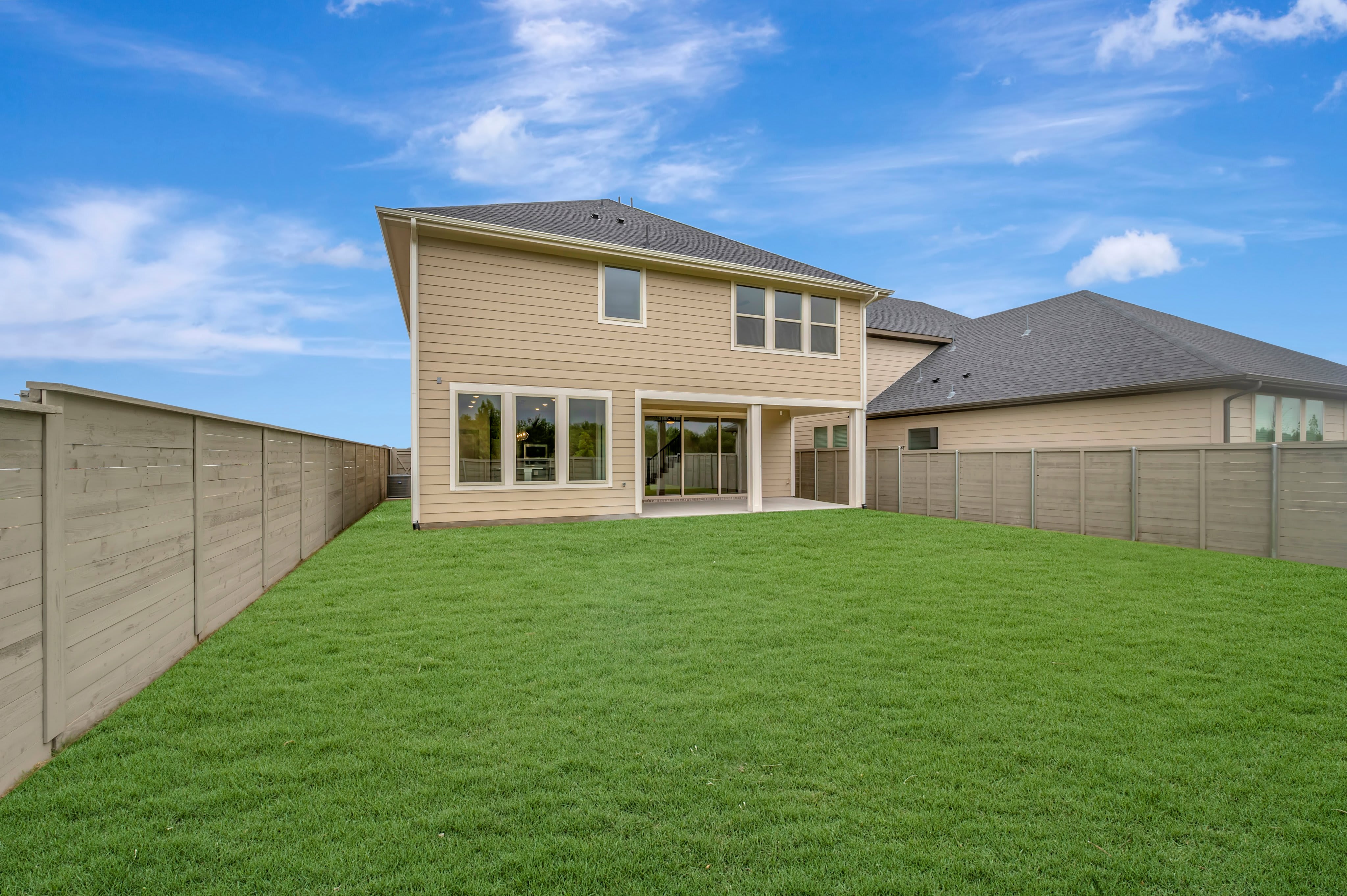A freshly mowed green lawn fills the foreground, leading to a tan-colored house with large windows and a covered patio in the background, all enclosed by a wooden fence under a bright blue sky with scattered clouds.