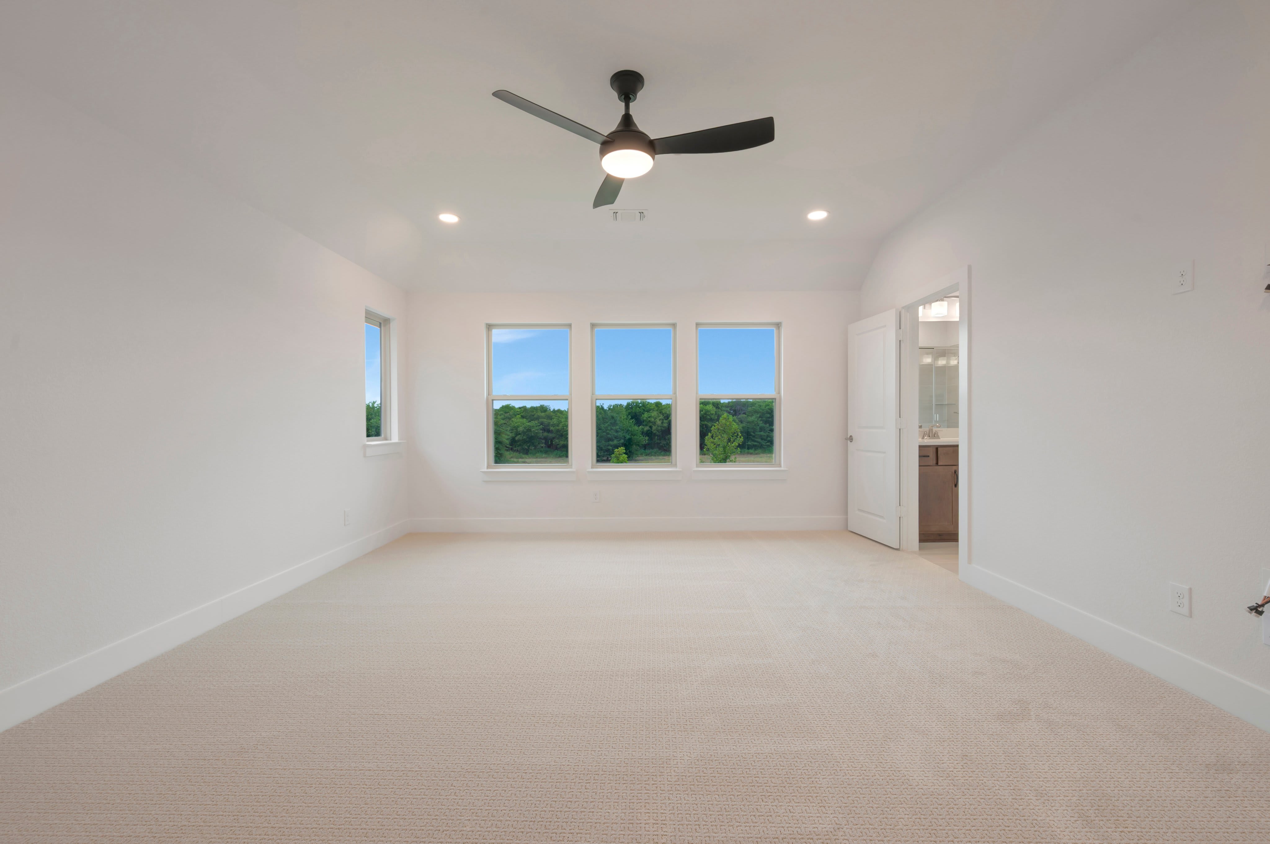 A spacious, carpeted room with a ceiling fan and three large windows overlooking a green landscape is in the foreground, while an open doorway reveals a glimpse of a bathroom in the background.
