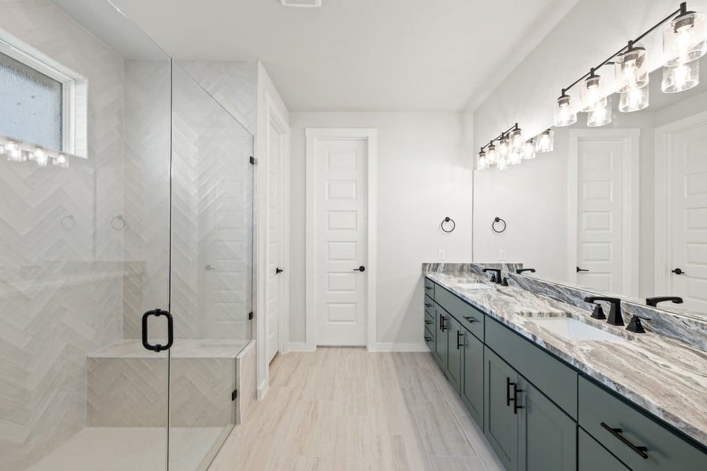 A modern bathroom features a glass-enclosed shower with a built-in bench in the foreground and a double vanity with a large mirror and multiple light fixtures in the background.