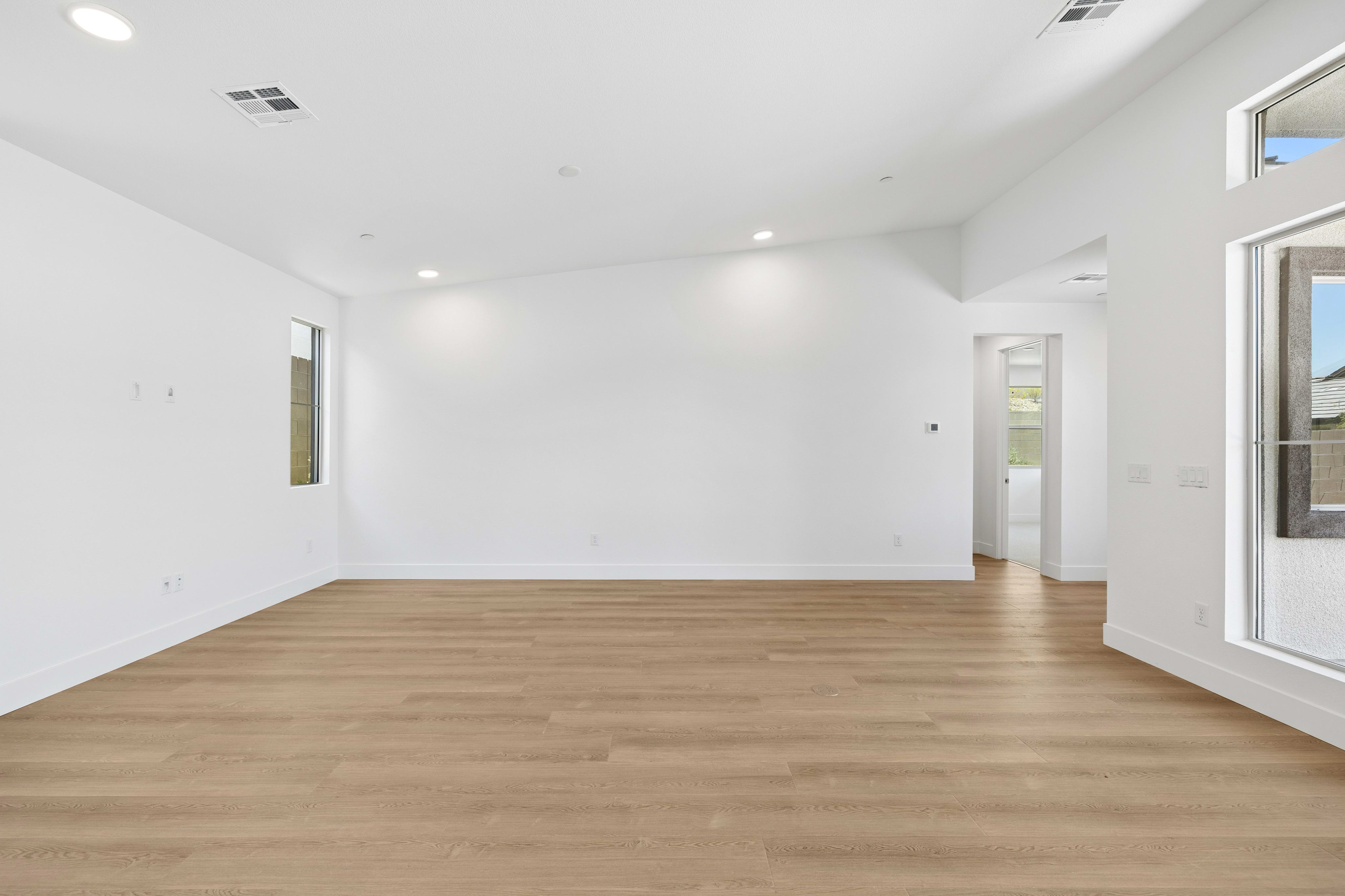 A spacious, empty room with light wood floors in the foreground opens to a hallway and an outdoor patio in the background.