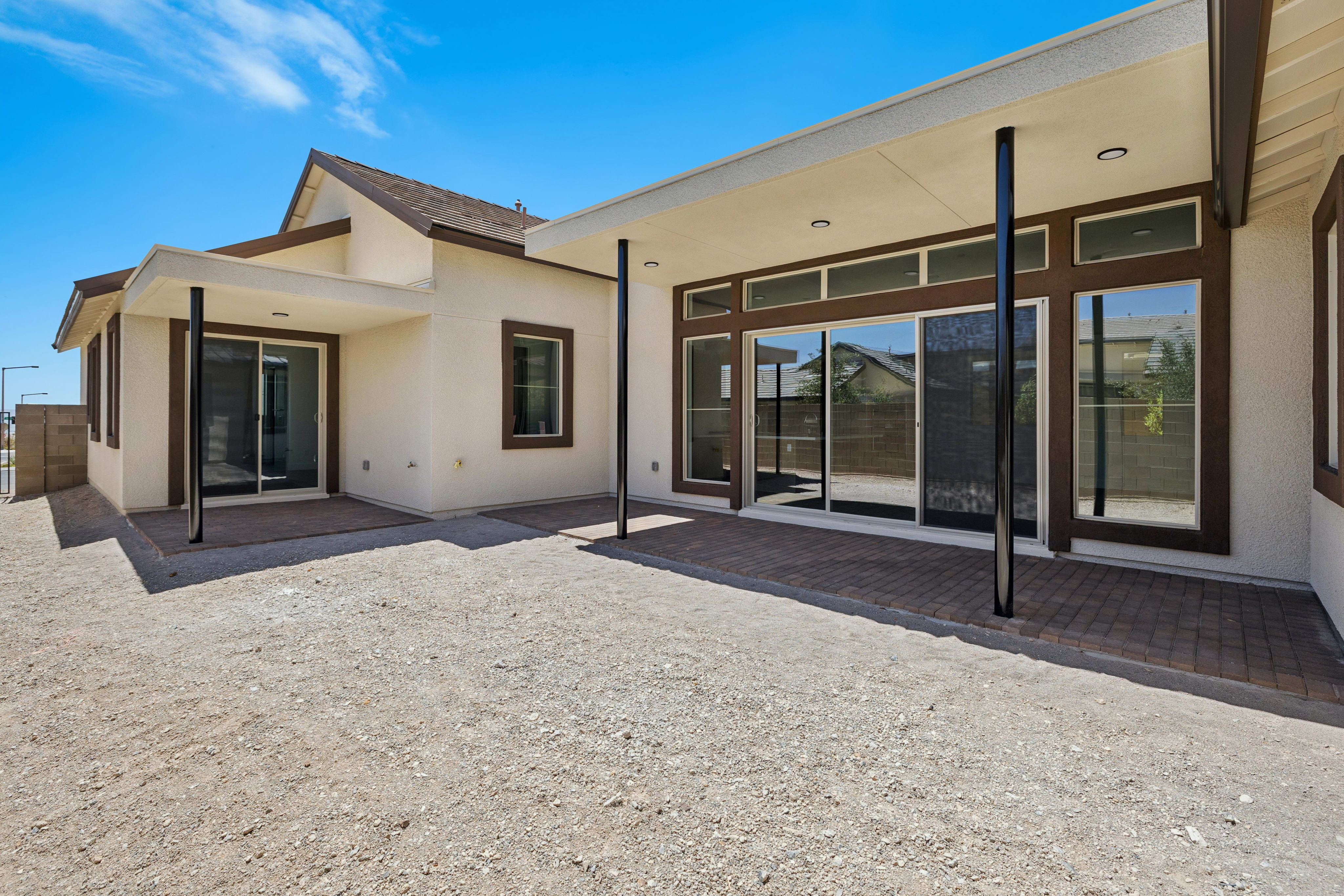 A gravel patio with brick accents leads to a modern house with large glass doors and windows, set against a bright blue sky.