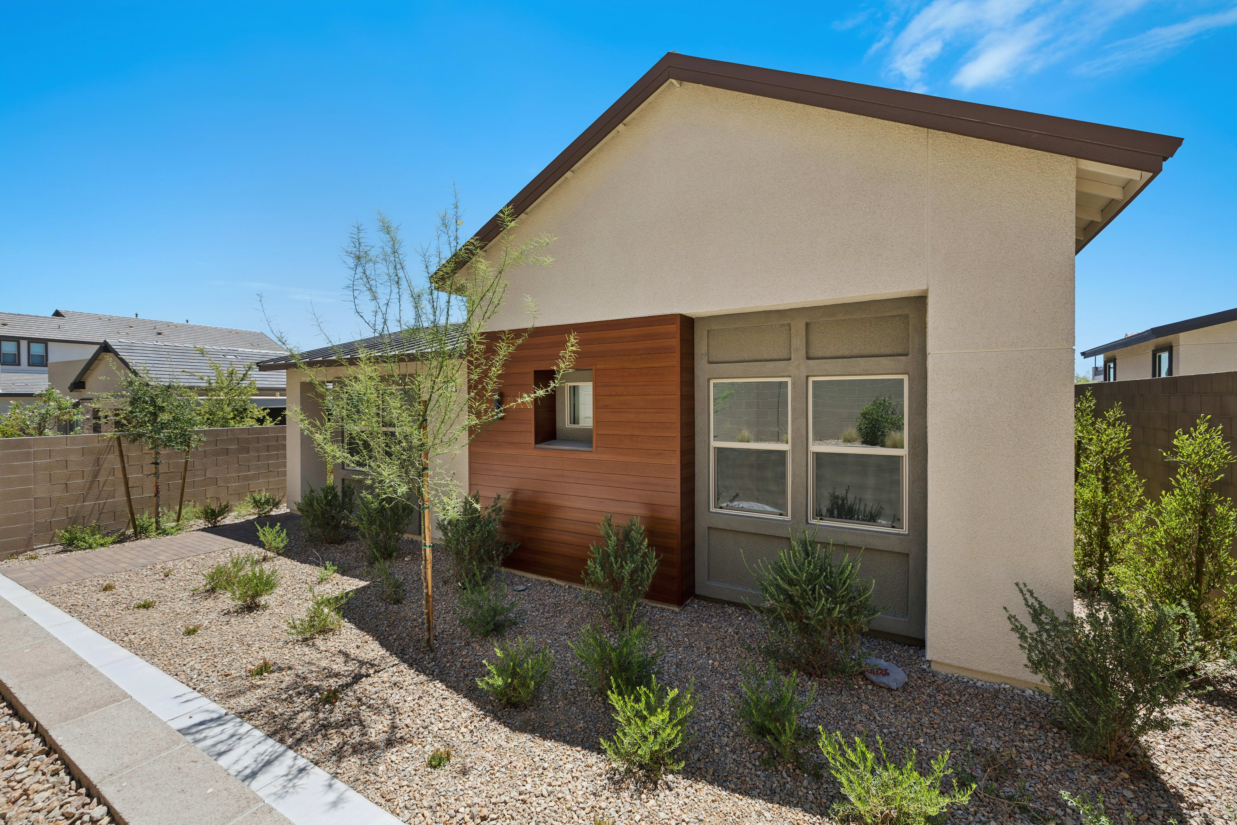 A modern stucco building with wood accents and a garage door sits in a xeriscaped yard with sparse green bushes and gravel, under a clear blue sky with distant houses.