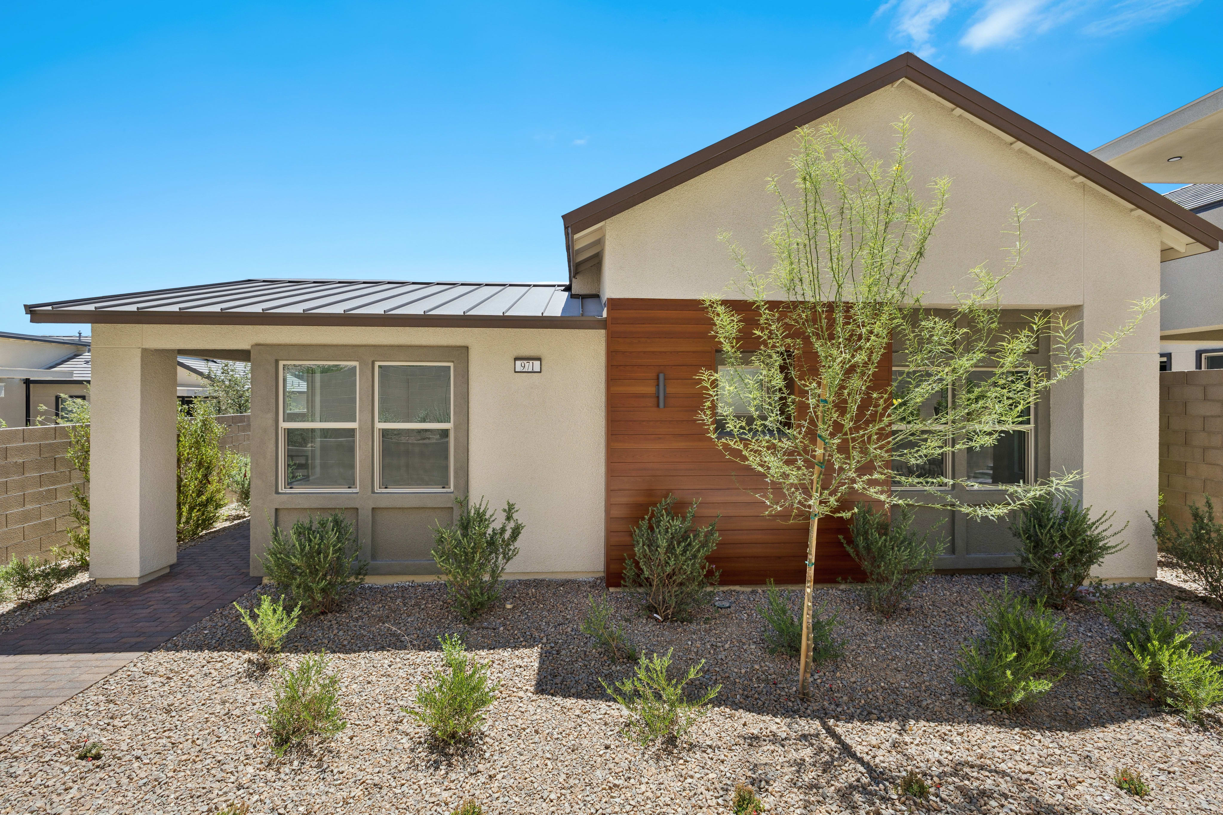 A modern stucco house with a metal roof and wood accents stands behind a gravel yard with low-lying bushes and a young tree, under a clear blue sky.