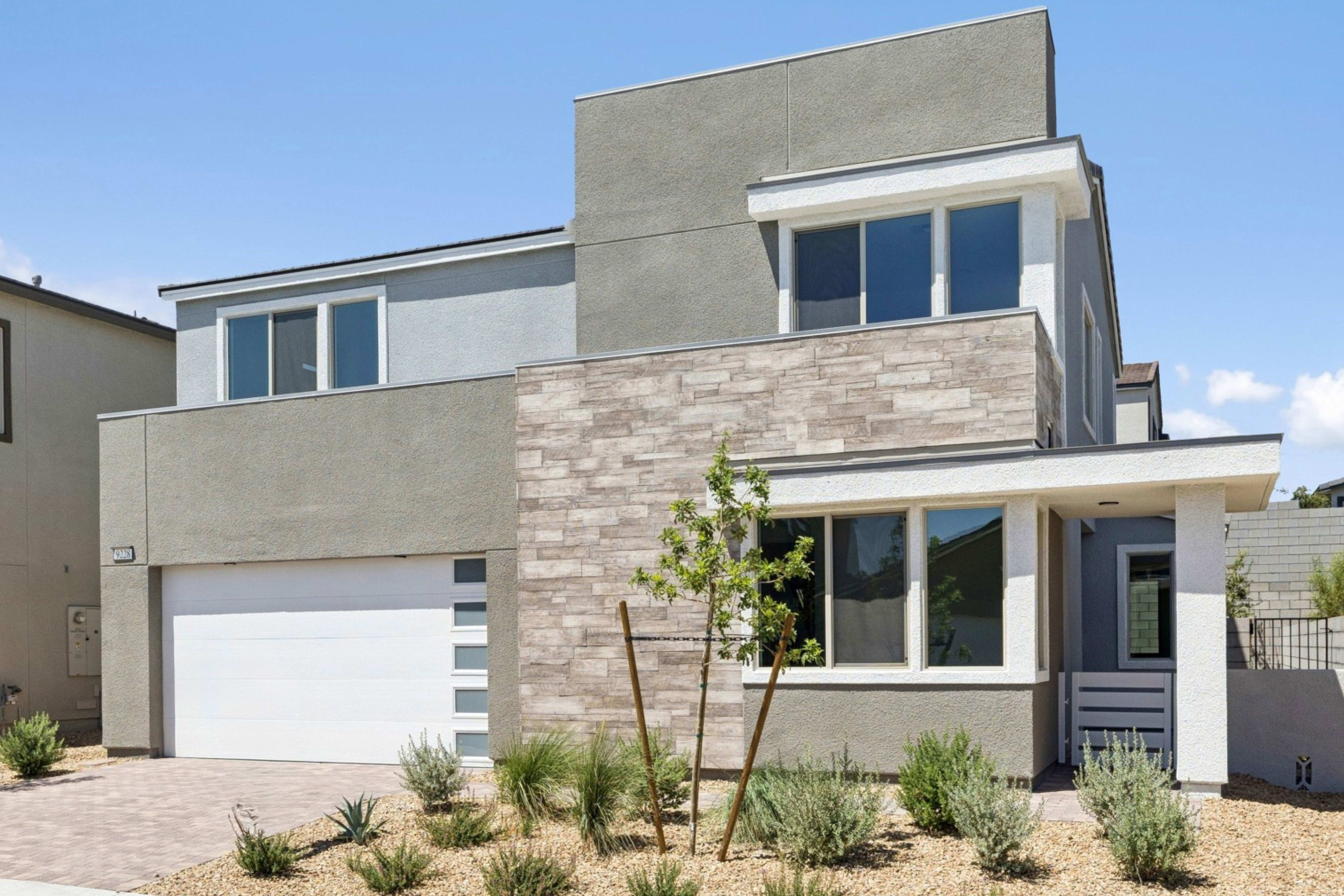 A modern gray house with a stone accent wall and a paved driveway sits in front of a clear blue sky.