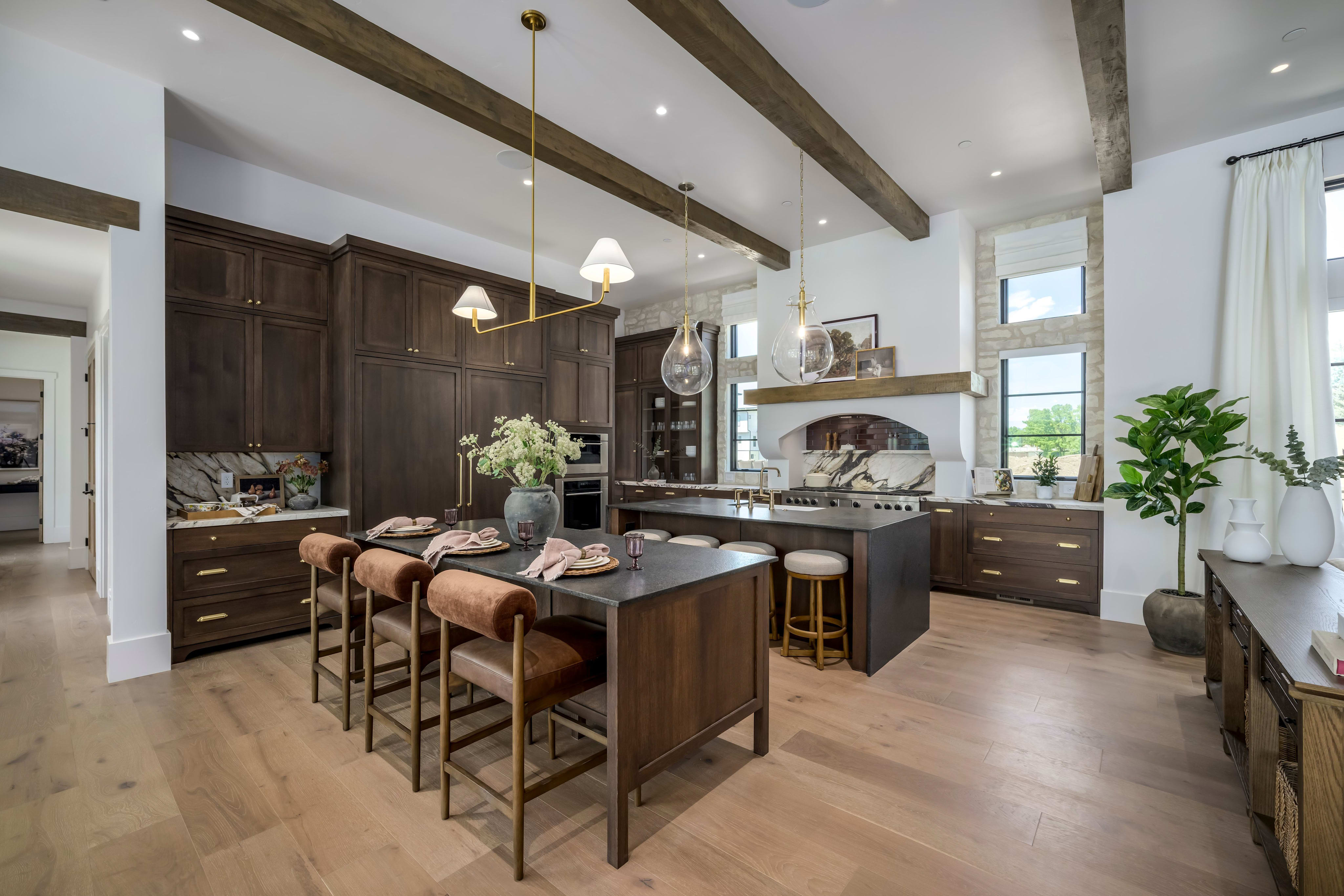 A spacious, modern kitchen with a dark wood island and seating in the foreground, and a large, well-equipped kitchen area with windows and exposed brick in the background.