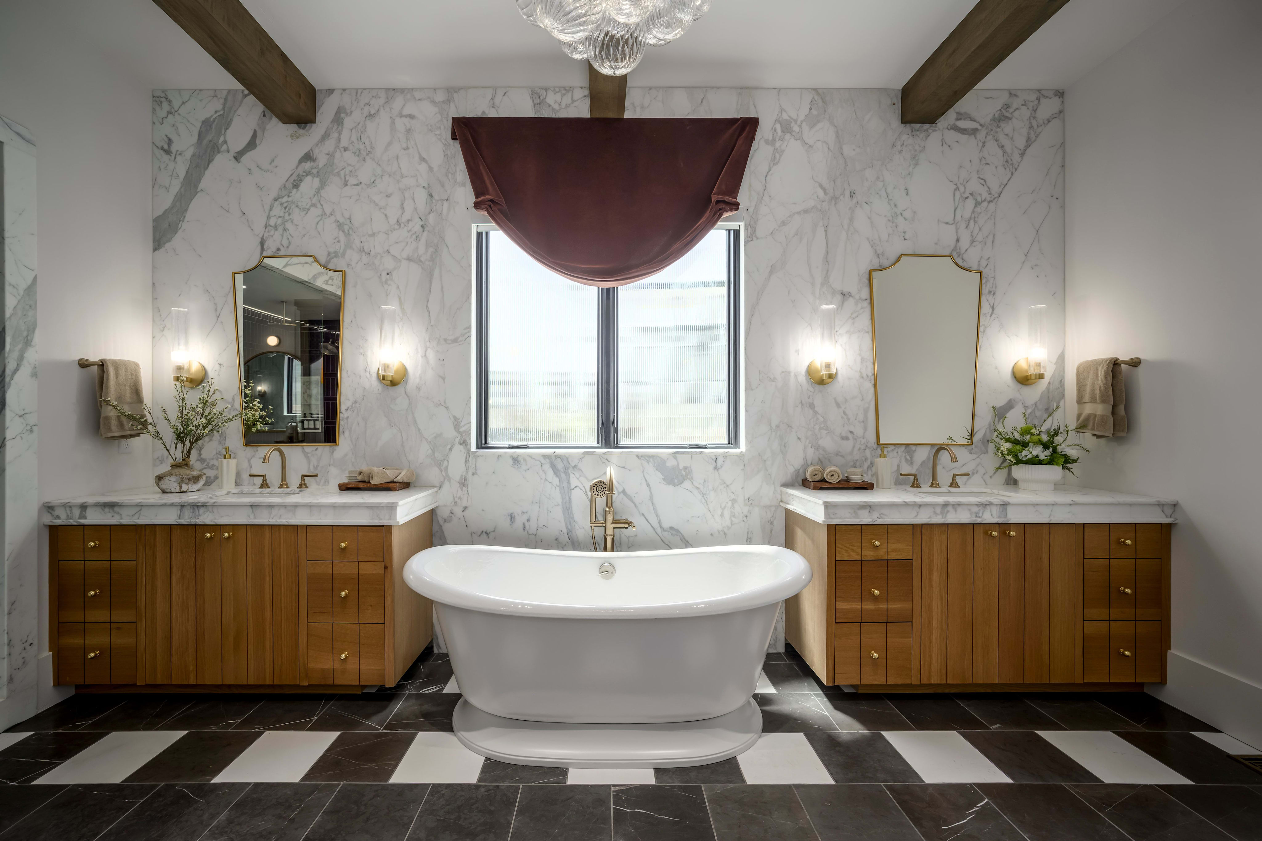 A freestanding white bathtub sits centered on a black and white tiled floor in front of a large window, flanked by dual vanities with marble countertops and wooden cabinets, all set against a marble-tiled wall with exposed wooden beams overhead.