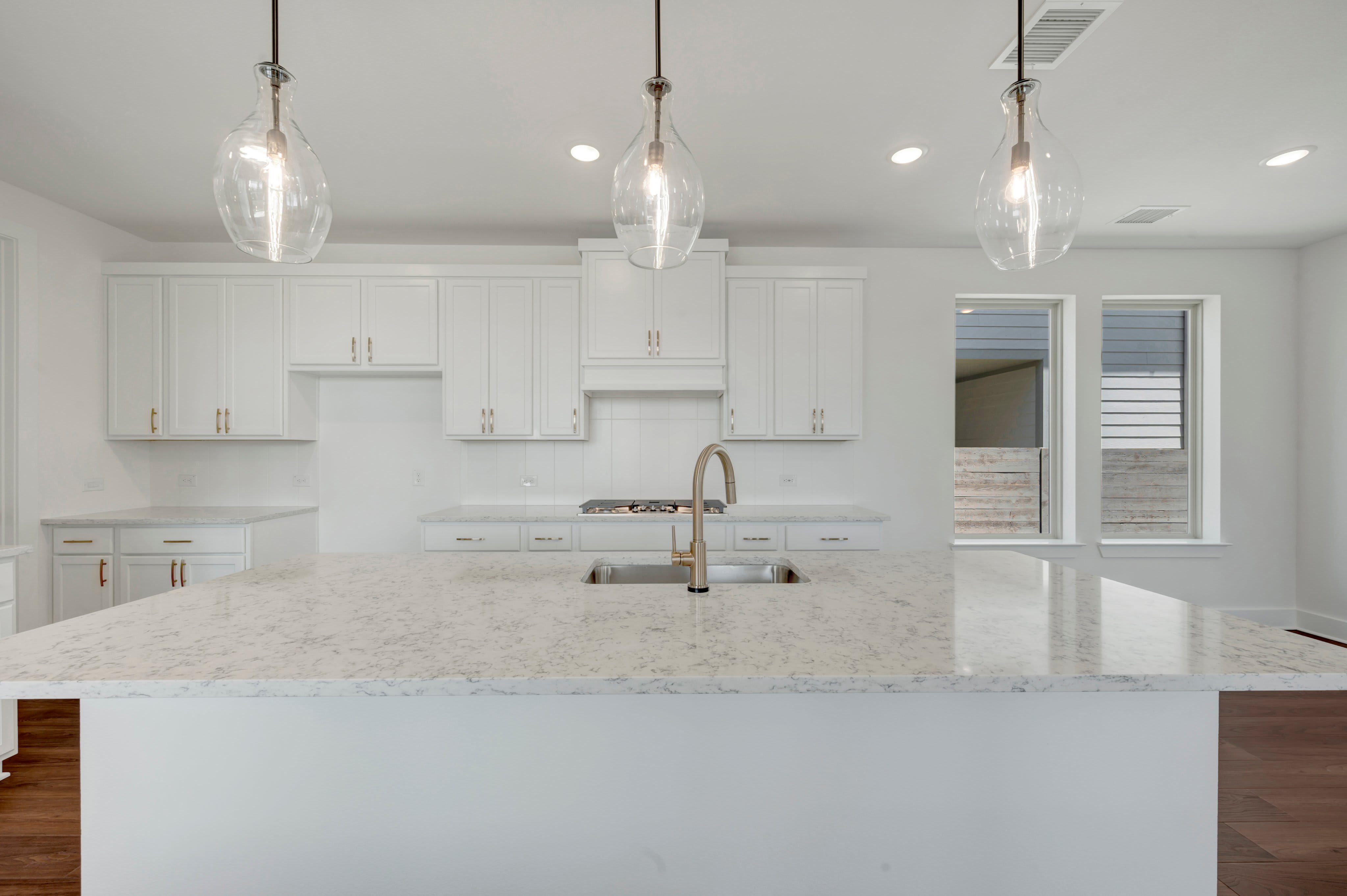 A modern white kitchen with a large island in the foreground and windows looking out to a backyard in the background.