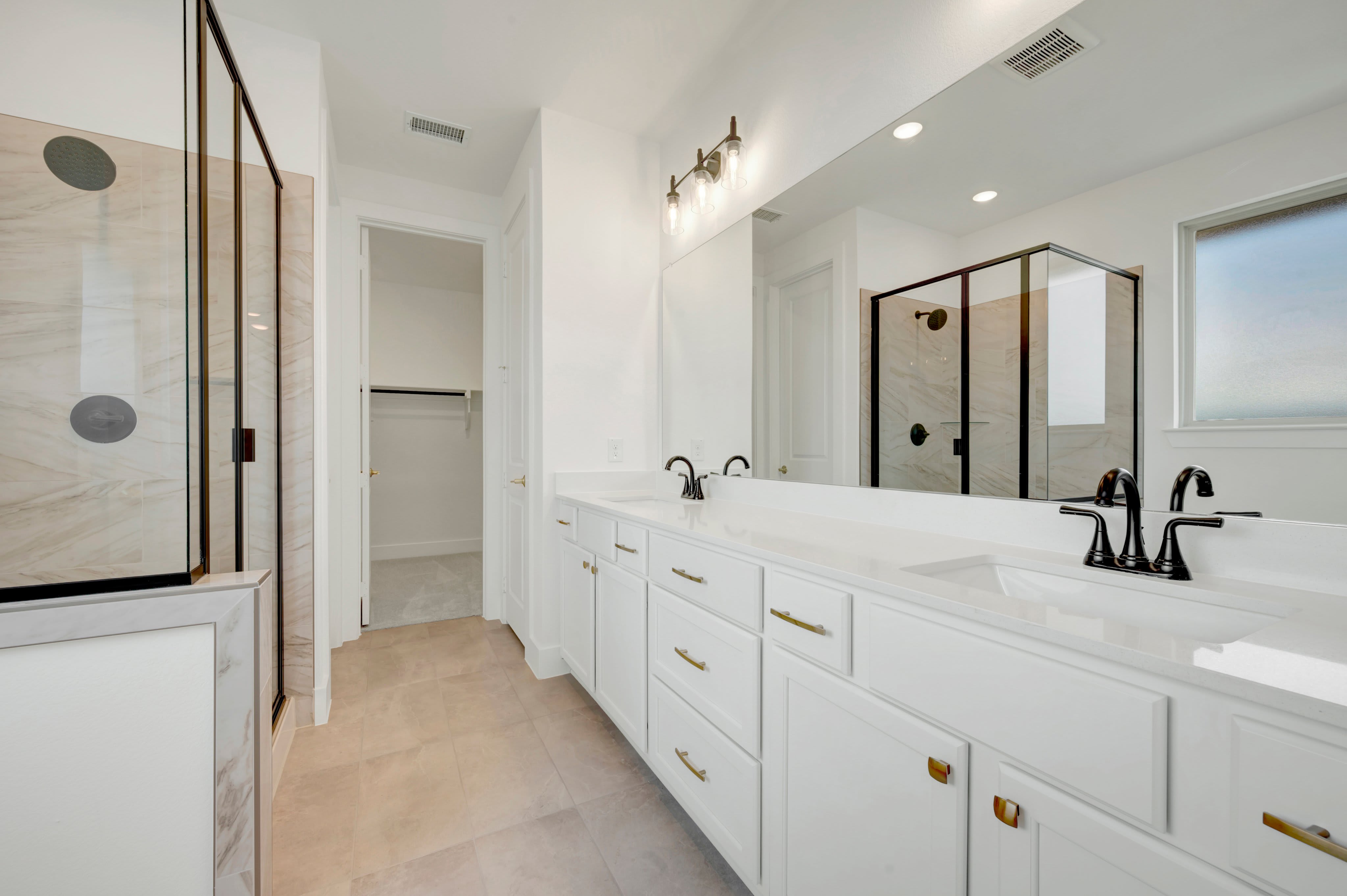 A modern white bathroom vanity with dual sinks and gold hardware is in the foreground, while a glass-enclosed shower and a walk-in closet are visible in the background.