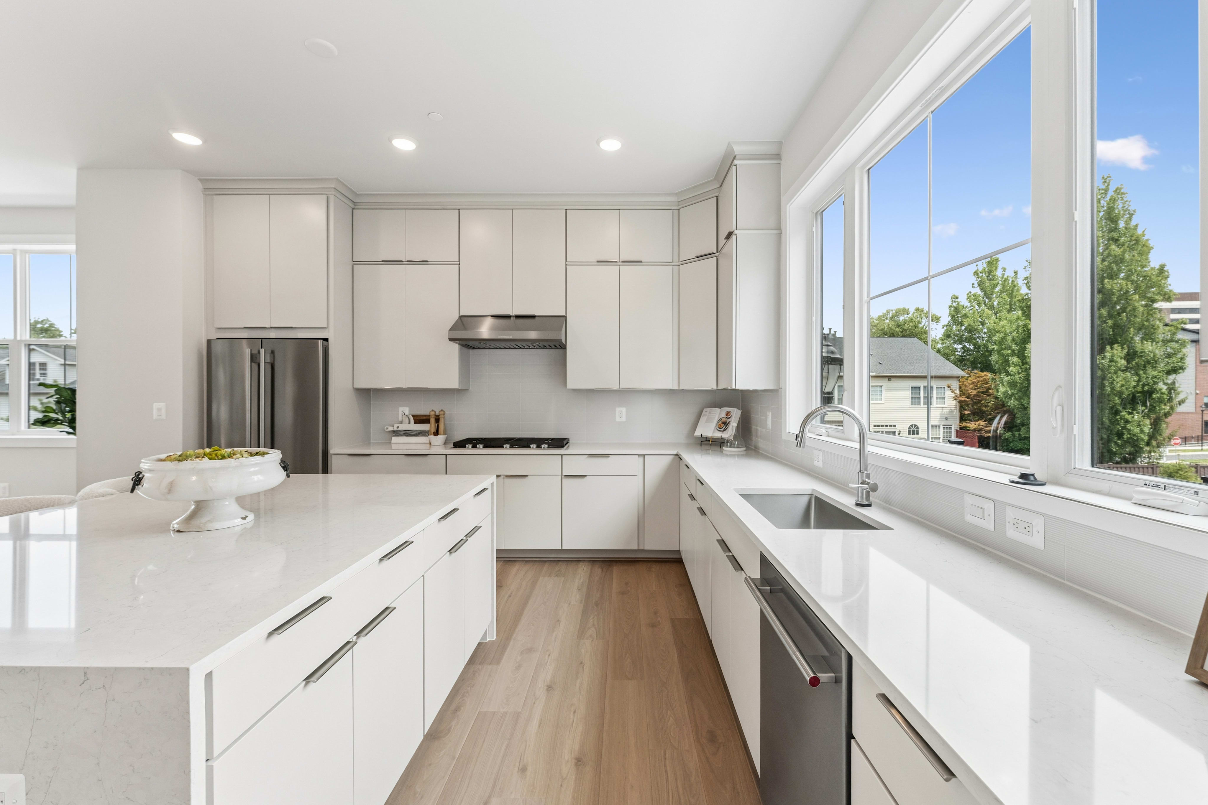 A modern white kitchen with a large island in the foreground and a bright window overlooking a suburban neighborhood in the background.