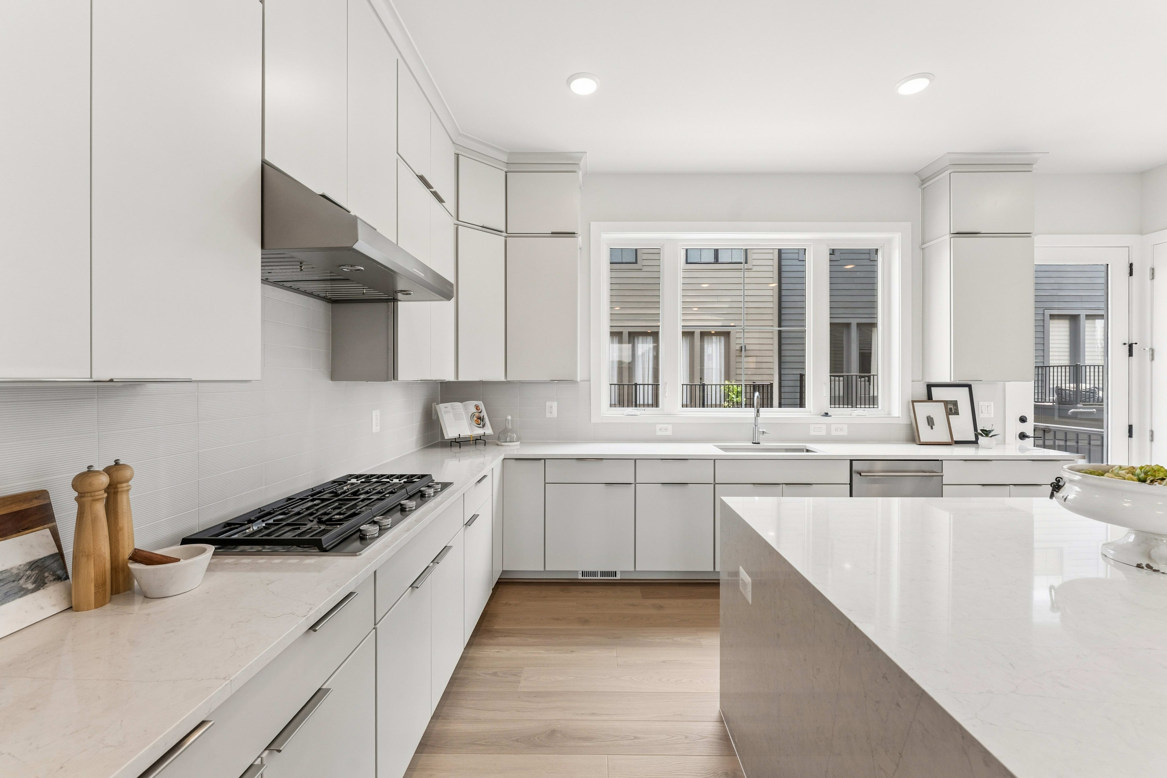 A modern white kitchen with a gas stove and island in the foreground, and a large window overlooking houses in the background.