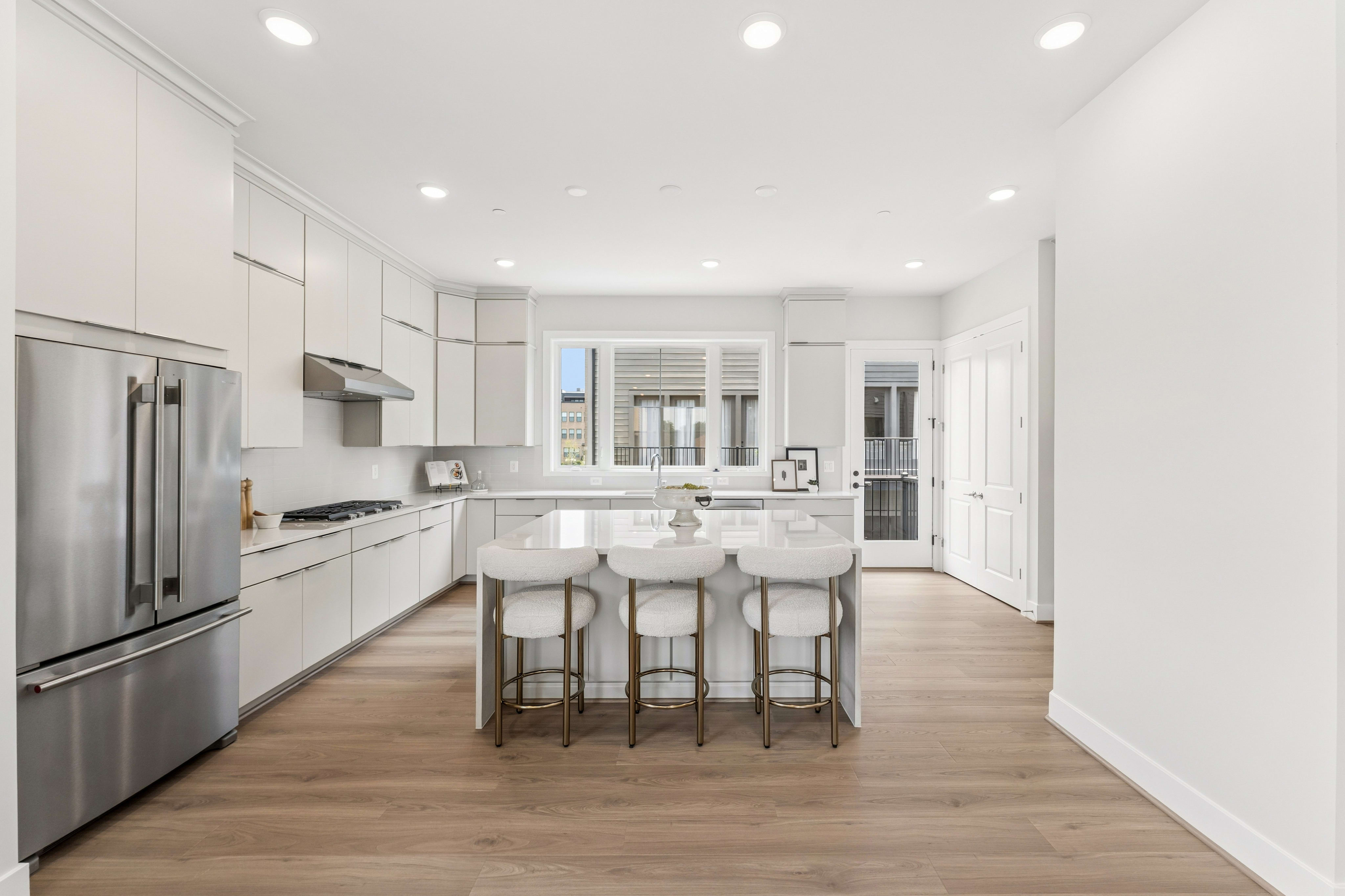 A modern white kitchen with a stainless steel refrigerator and island with bar stools in the foreground, and a bright window and doorway in the background.