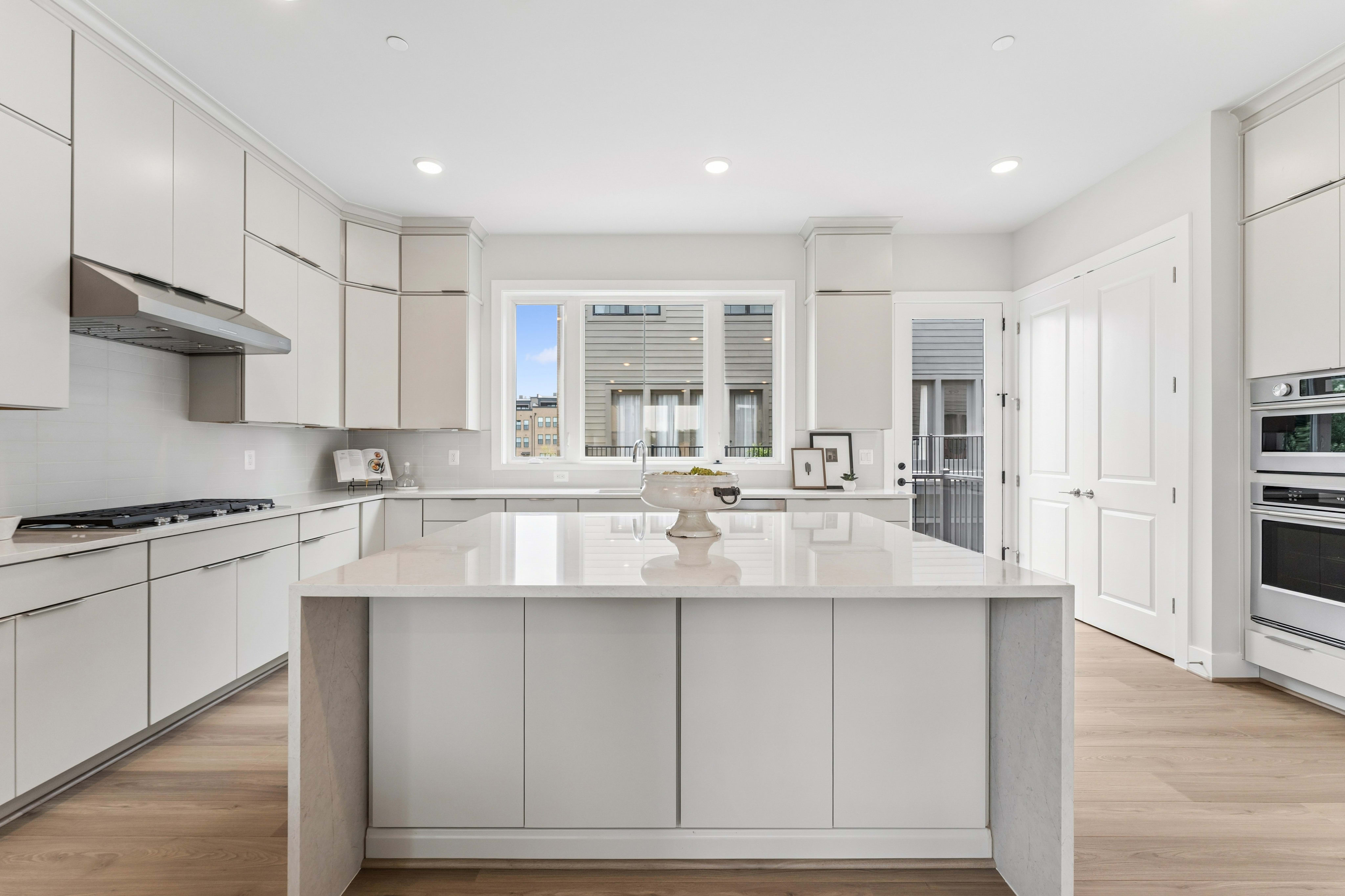 A modern white kitchen with a large island in the foreground and a window overlooking houses in the background.