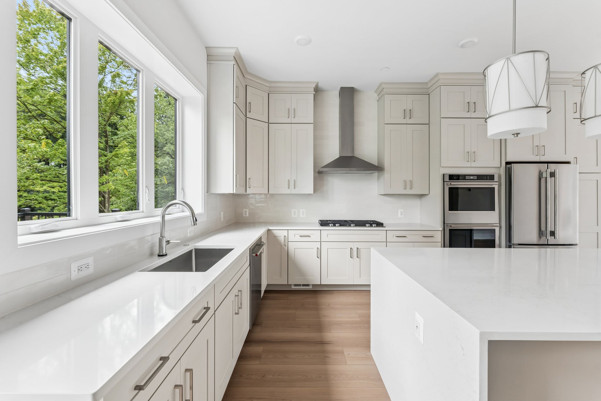 A modern white kitchen with a large island in the foreground and a window overlooking lush green trees in the background.