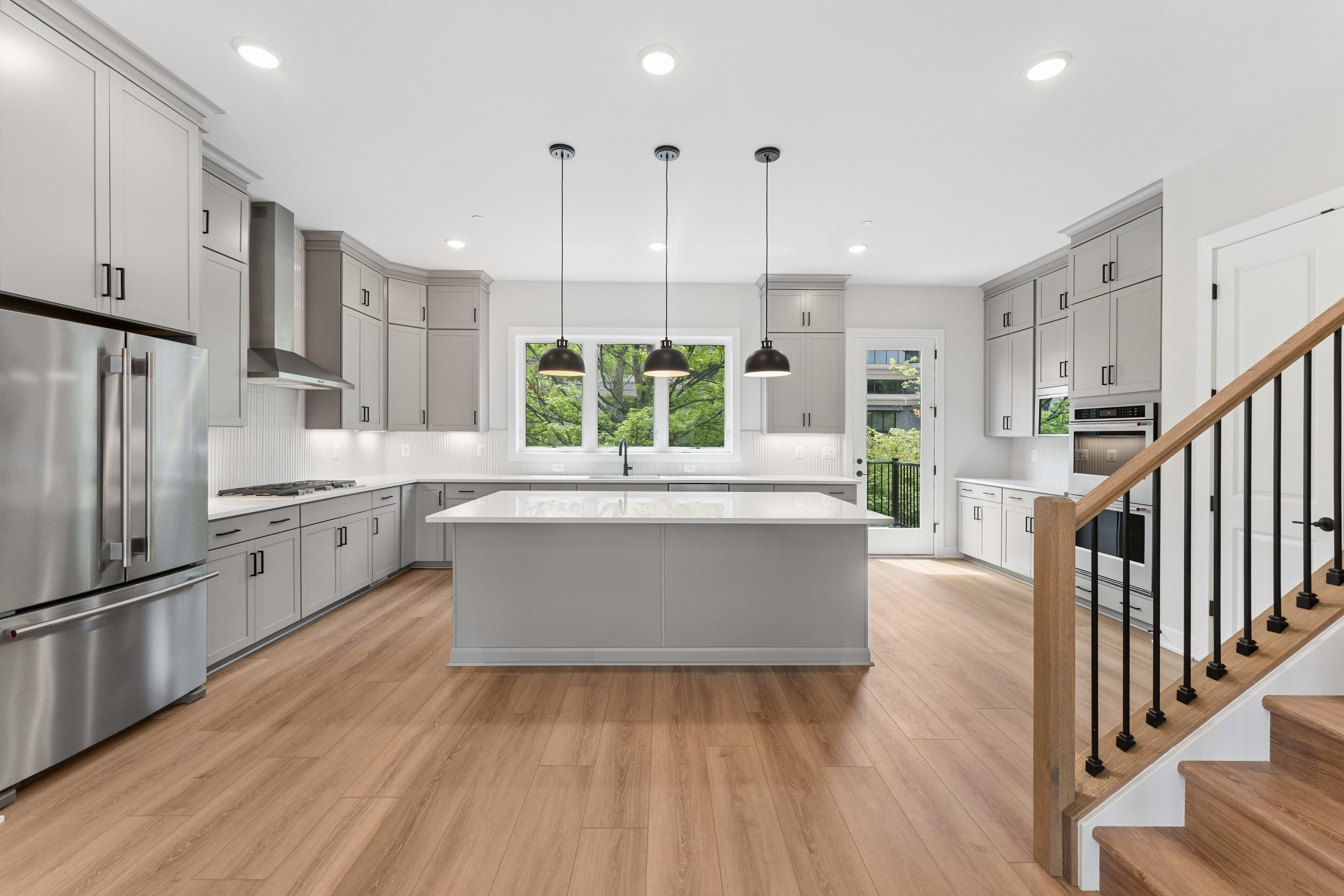 A modern gray kitchen with a large island in the foreground and a staircase leading upstairs in the background.