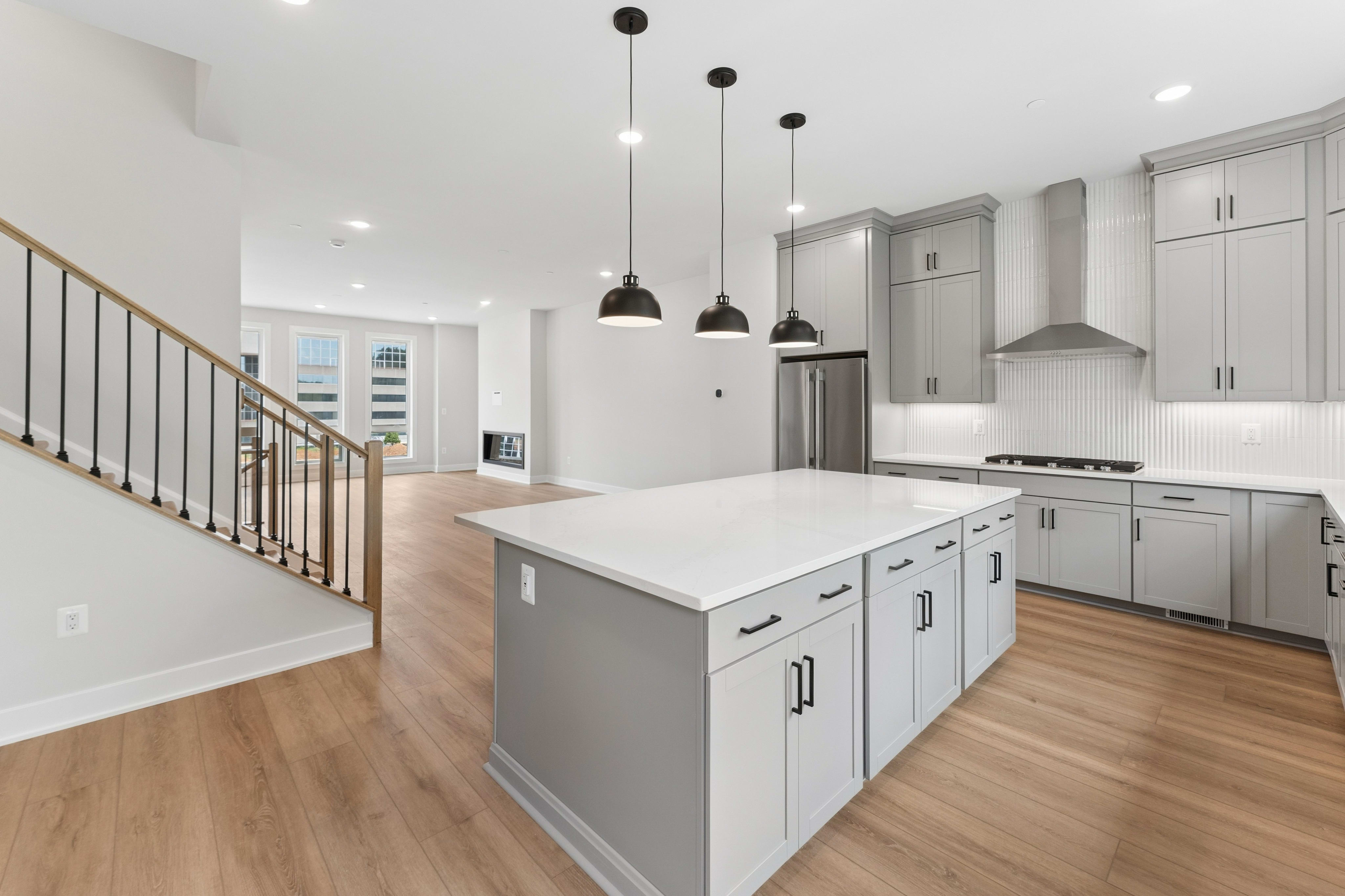 A modern kitchen island with a white countertop sits in the foreground, while a spacious living area with a fireplace and staircase is visible in the background.