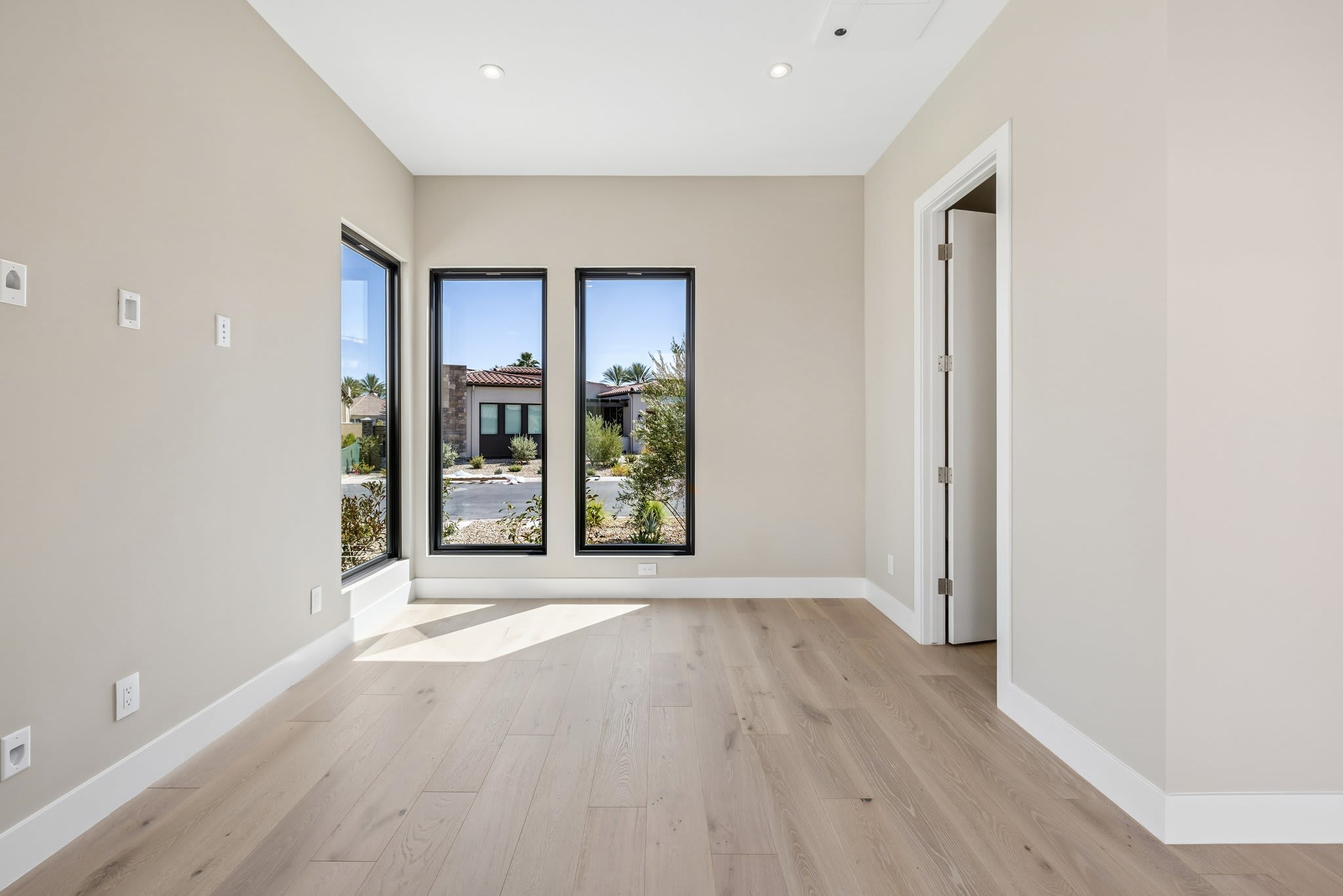 A bright, empty room with light wood floors in the foreground opens to a view of a suburban street through three large windows in the background.