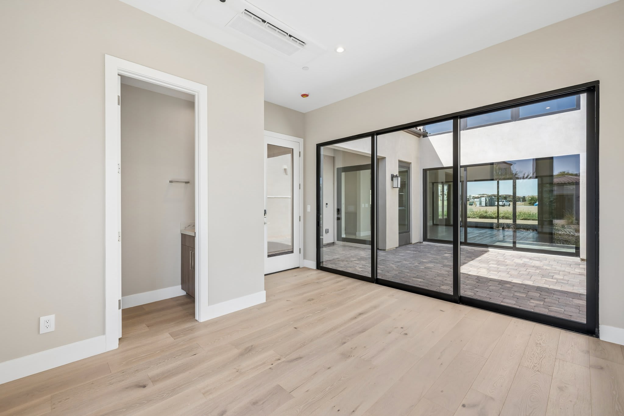 A bright, modern room with light wood floors in the foreground opens to a courtyard with a pool and modern architecture visible through large glass doors in the background.