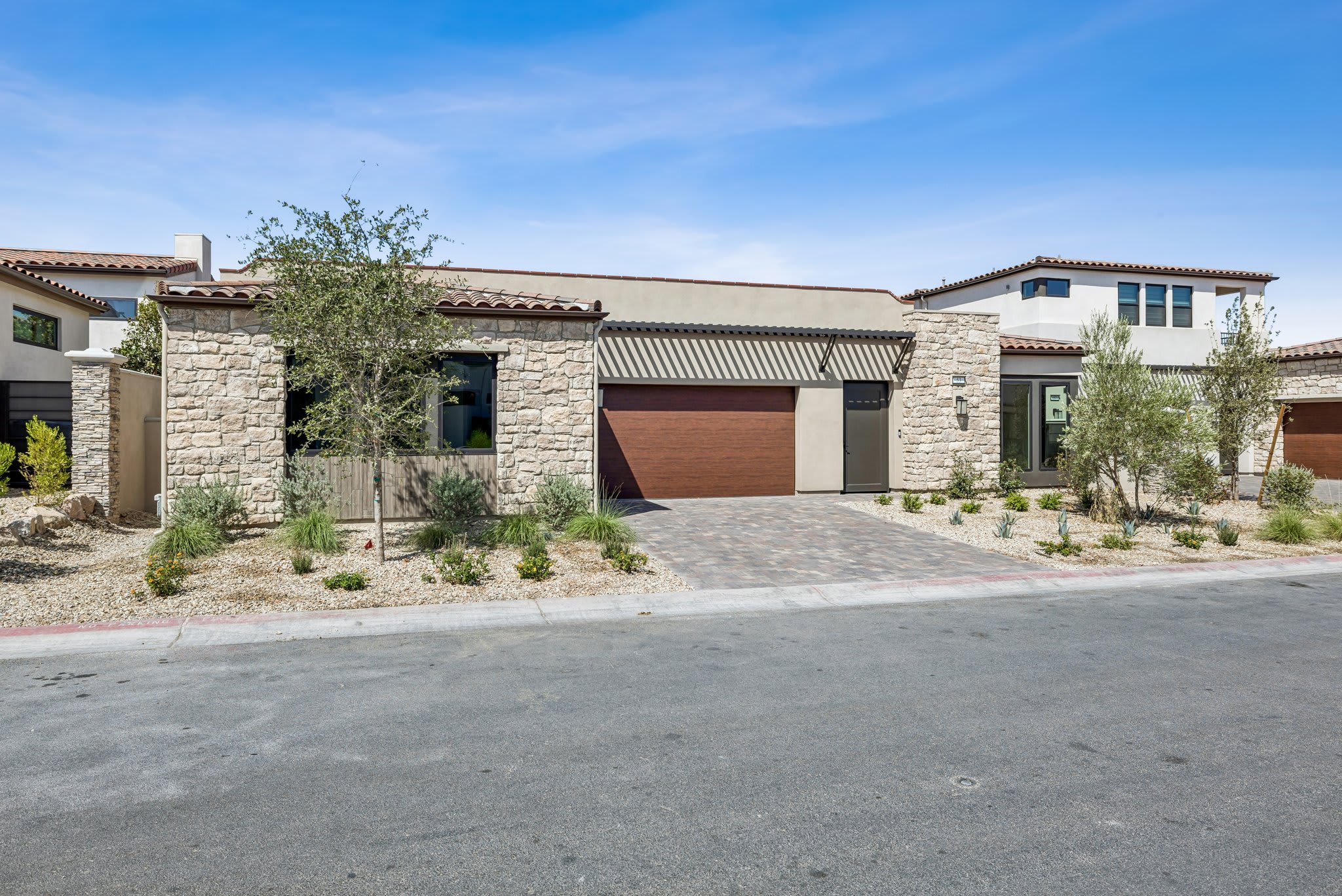 A modern stone-clad house with a brown garage door sits on a paved driveway in the foreground, while a clear blue sky with scattered clouds forms the background.