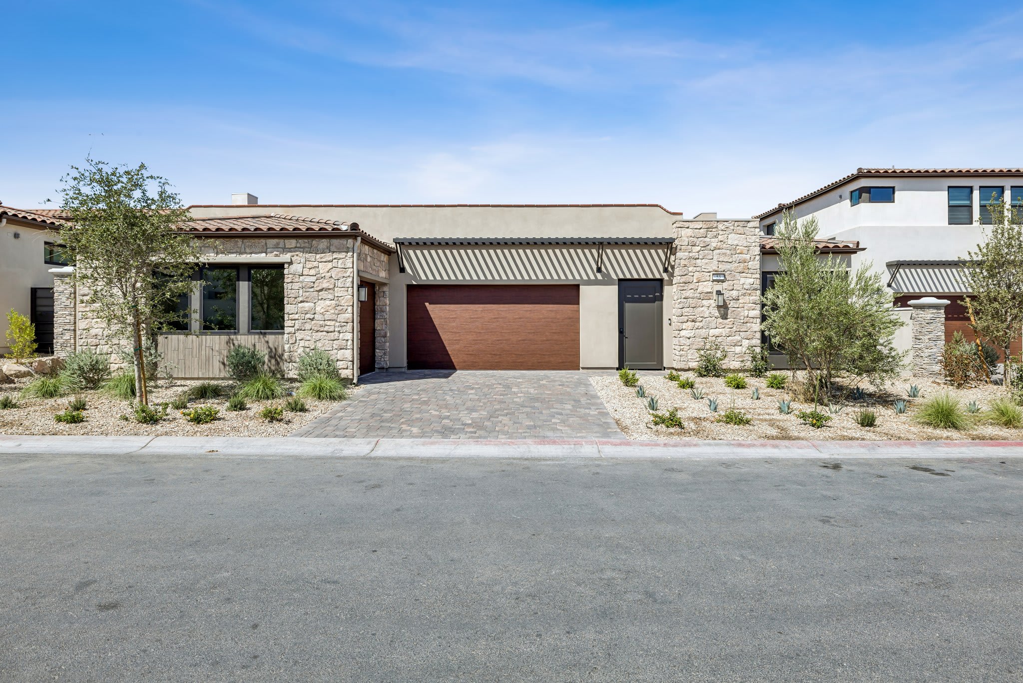 A modern stone-clad house with a brown garage door sits on a paved driveway in the foreground, while a clear blue sky with a few clouds forms the background.