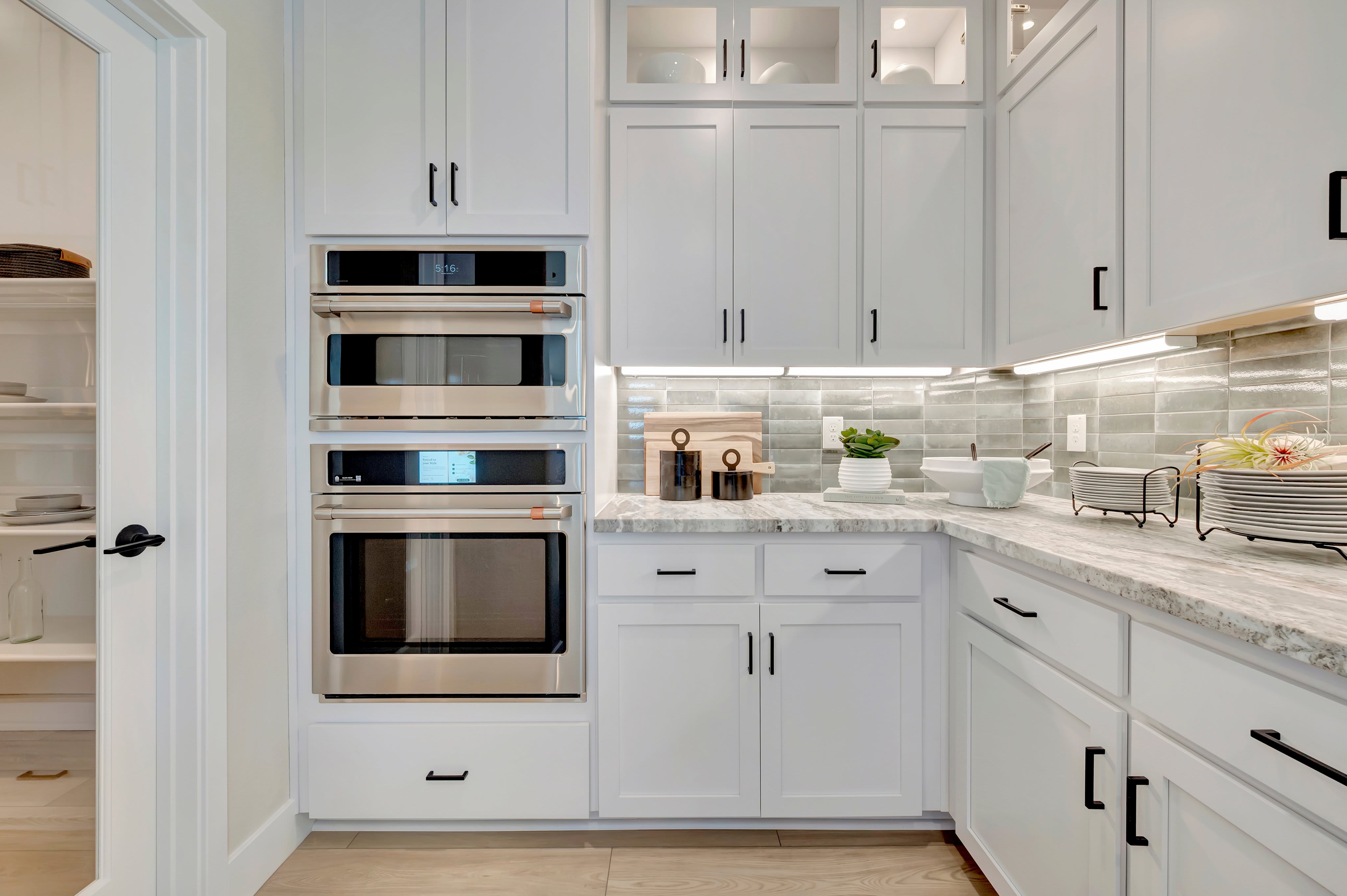 A modern white kitchen with stainless steel double ovens in the foreground and a pantry with shelves in the background.