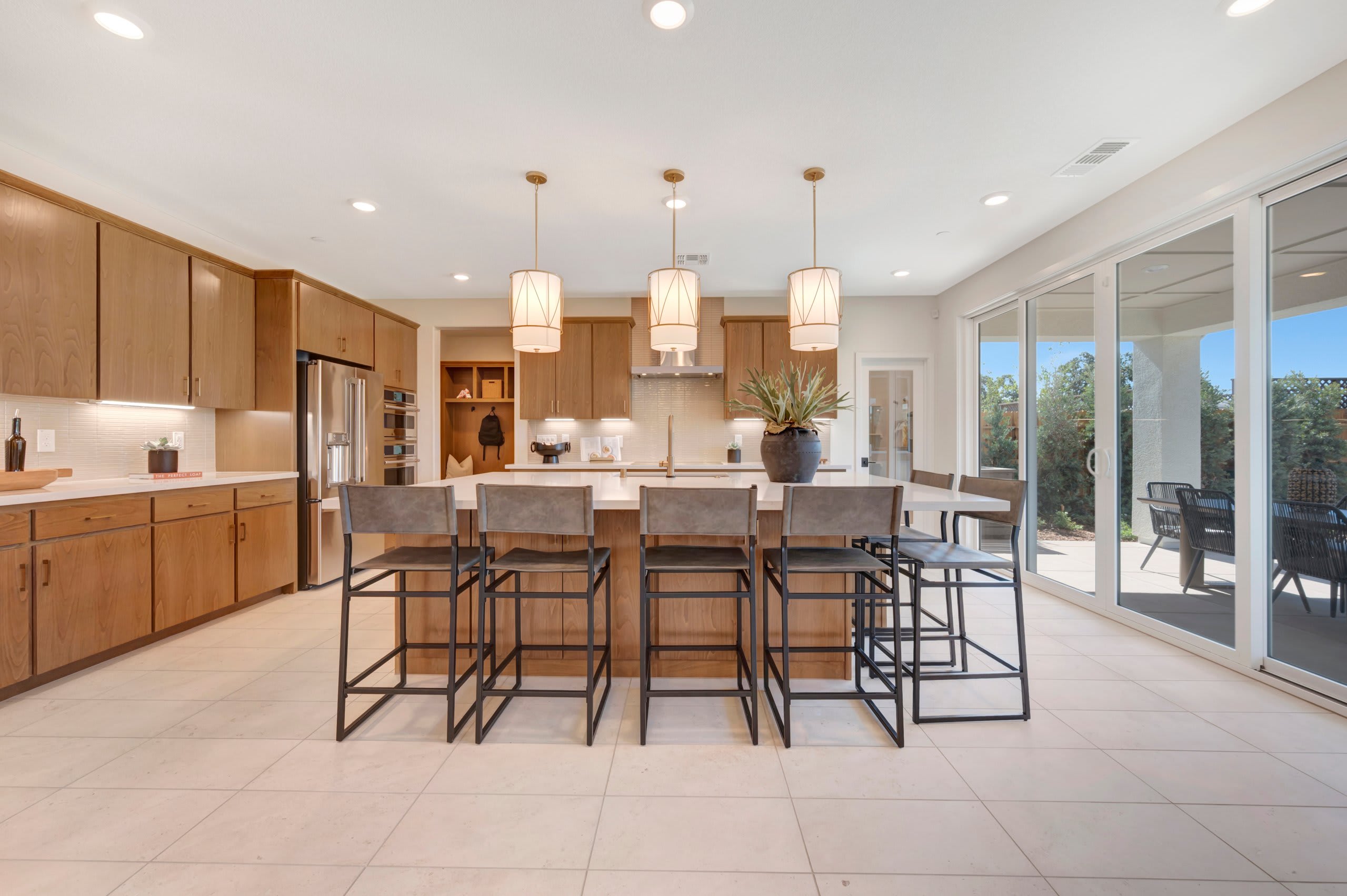 A modern kitchen with a large island and bar stools in the foreground opens to a patio with outdoor seating in the background.