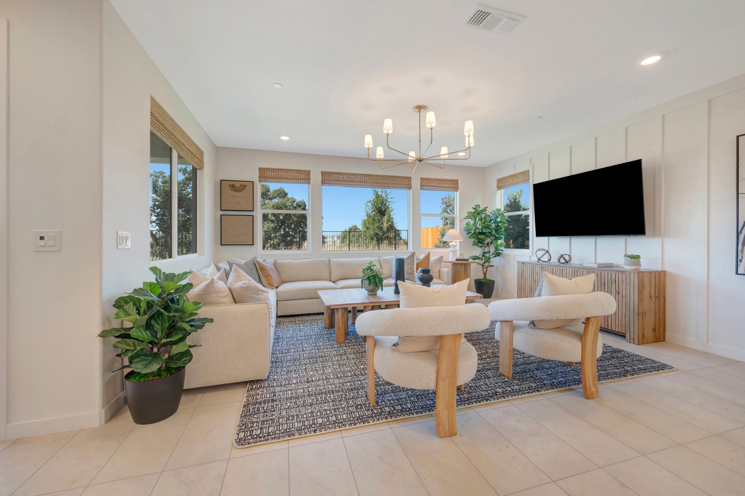 Two cream-colored armchairs sit on a patterned rug in the foreground, facing a large sectional sofa and a television mounted on a paneled wall in the background, with large windows offering a view of trees and a blue sky.