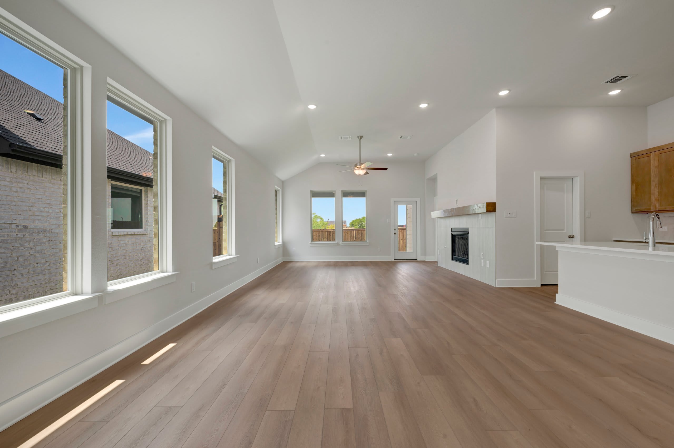 A spacious, modern living room with light wood floors in the foreground opens to a bright, airy space with large windows and a fireplace in the background.