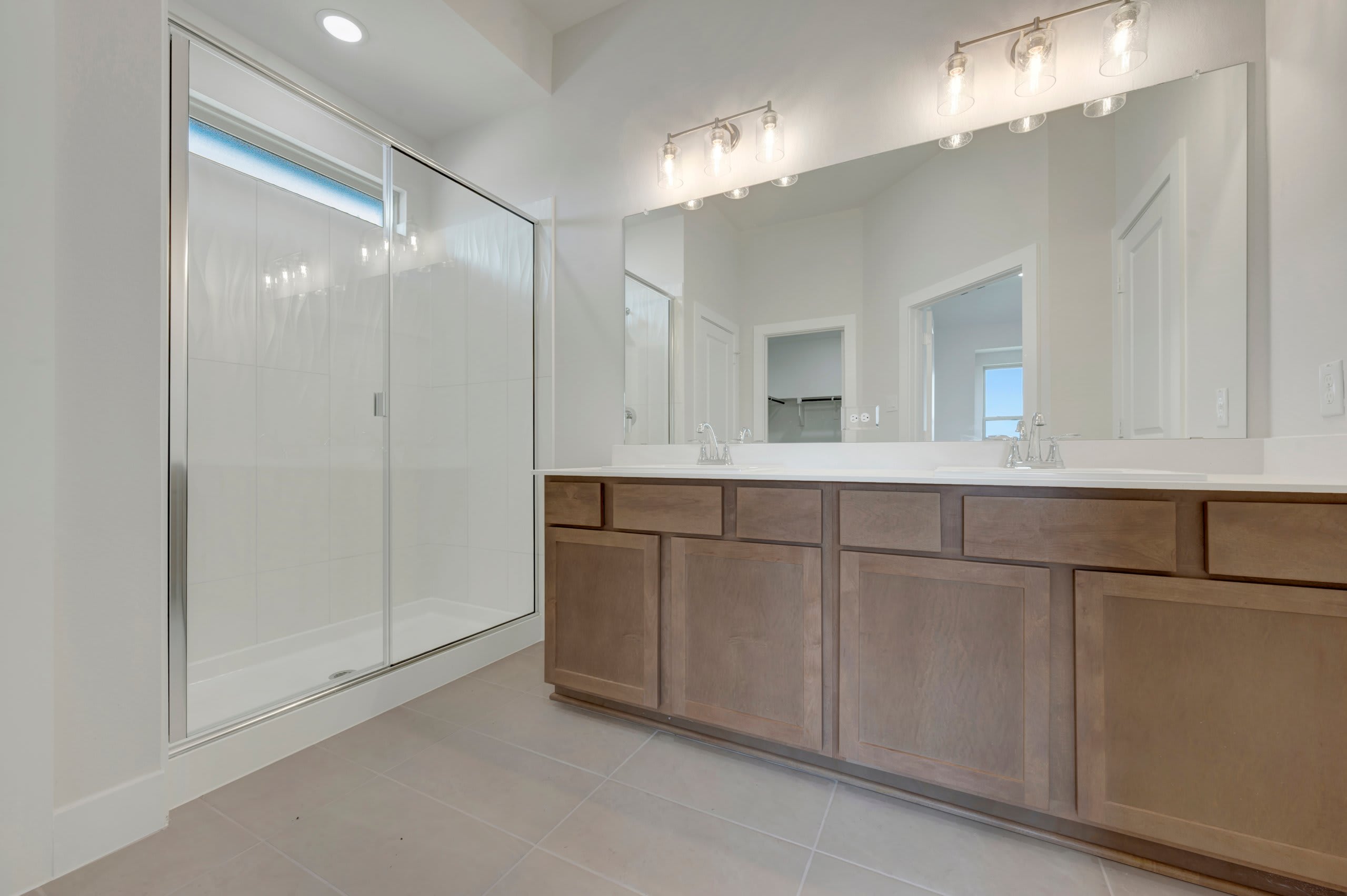 A modern bathroom features a glass-enclosed shower in the foreground and a double vanity with a large mirror reflecting the room in the background.