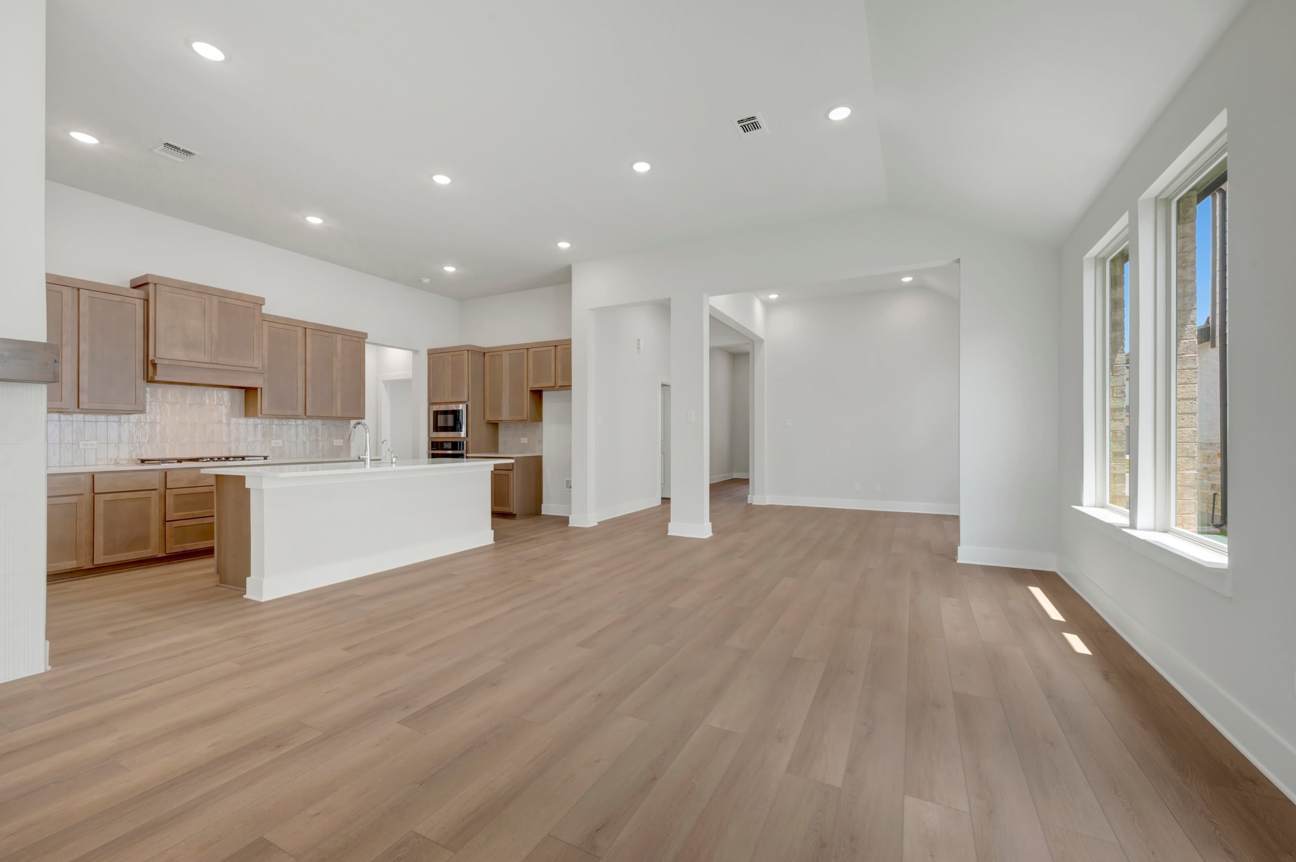 A spacious, modern living area with light wood flooring in the foreground opens to a kitchen with light wood cabinets and a white island, while the background reveals a bright, airy room with large windows.