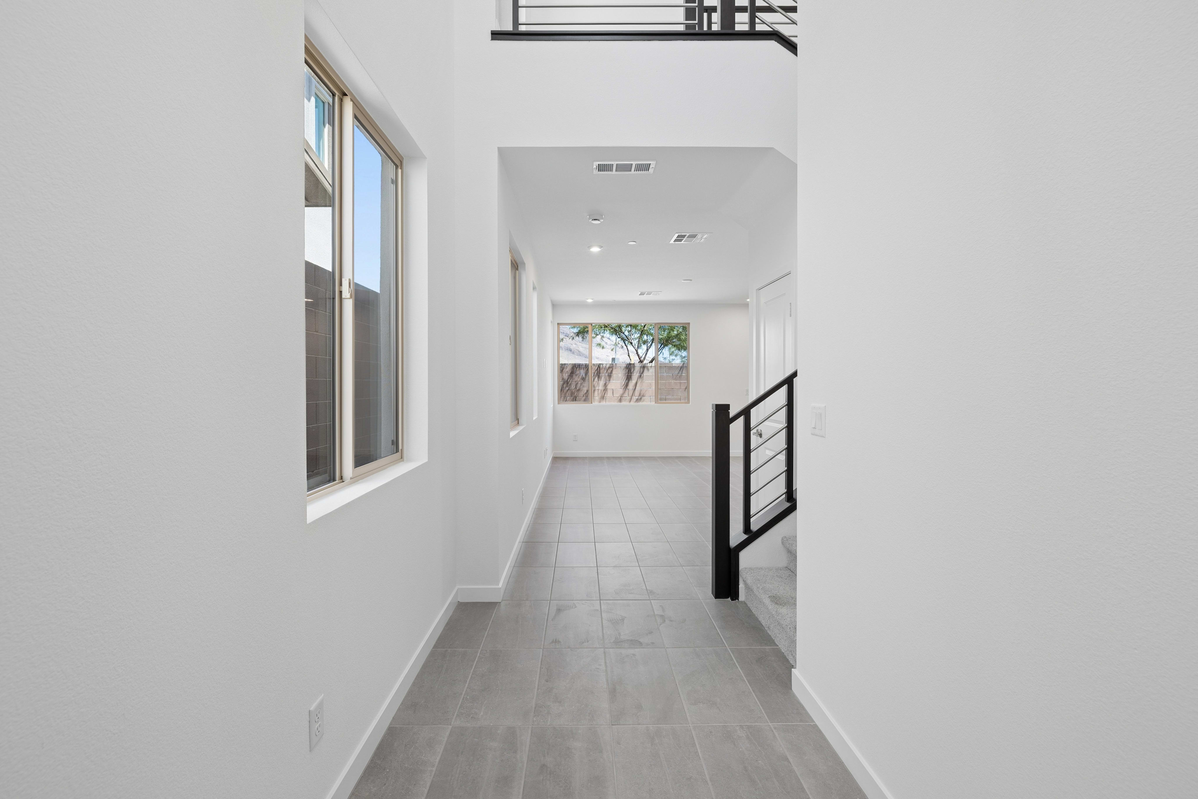 A modern hallway with tiled floors and large windows leads to a staircase and a room with a view of trees.