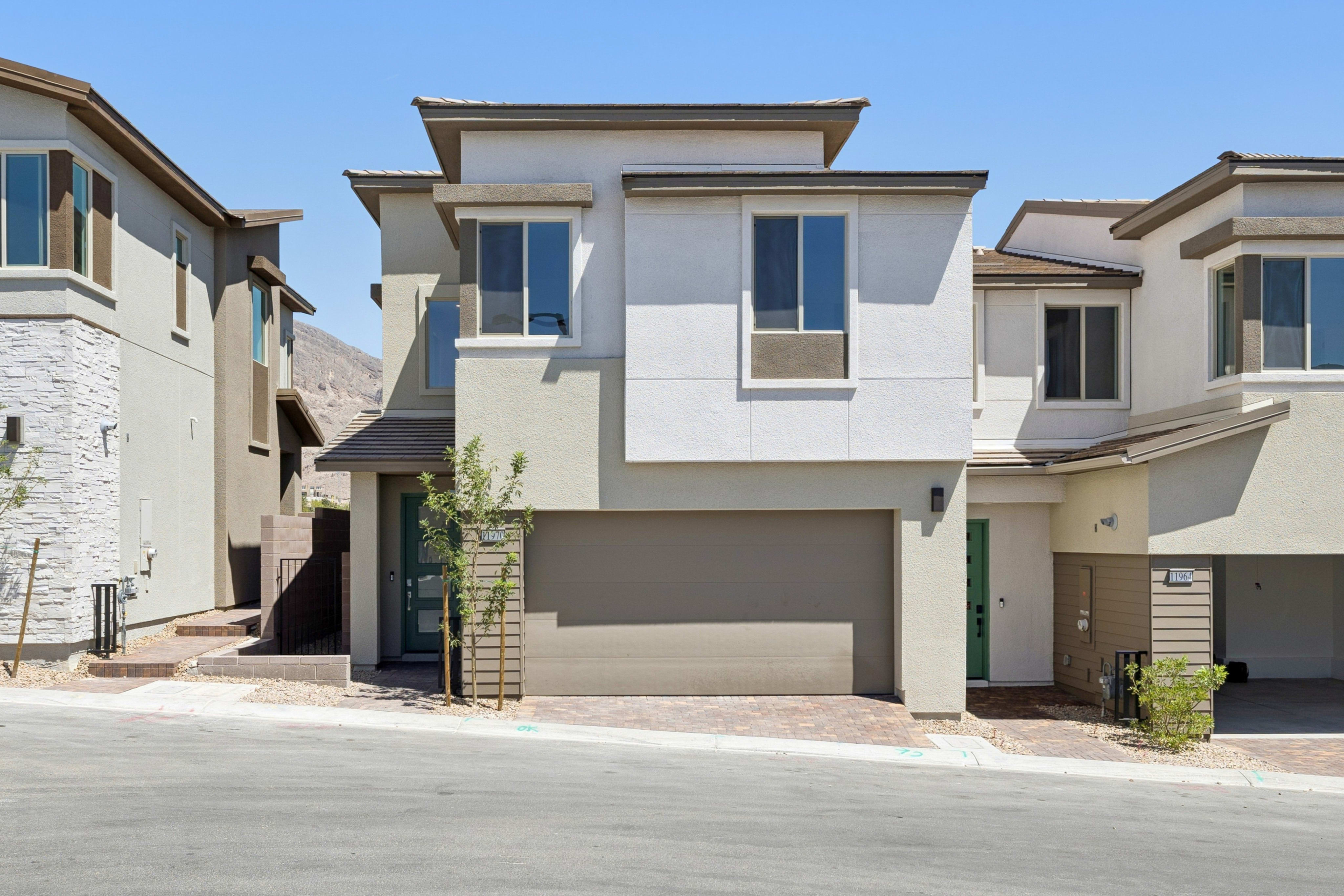 A modern townhouse with a beige and white exterior and a garage door is in the foreground, while a clear blue sky and distant hills form the background.