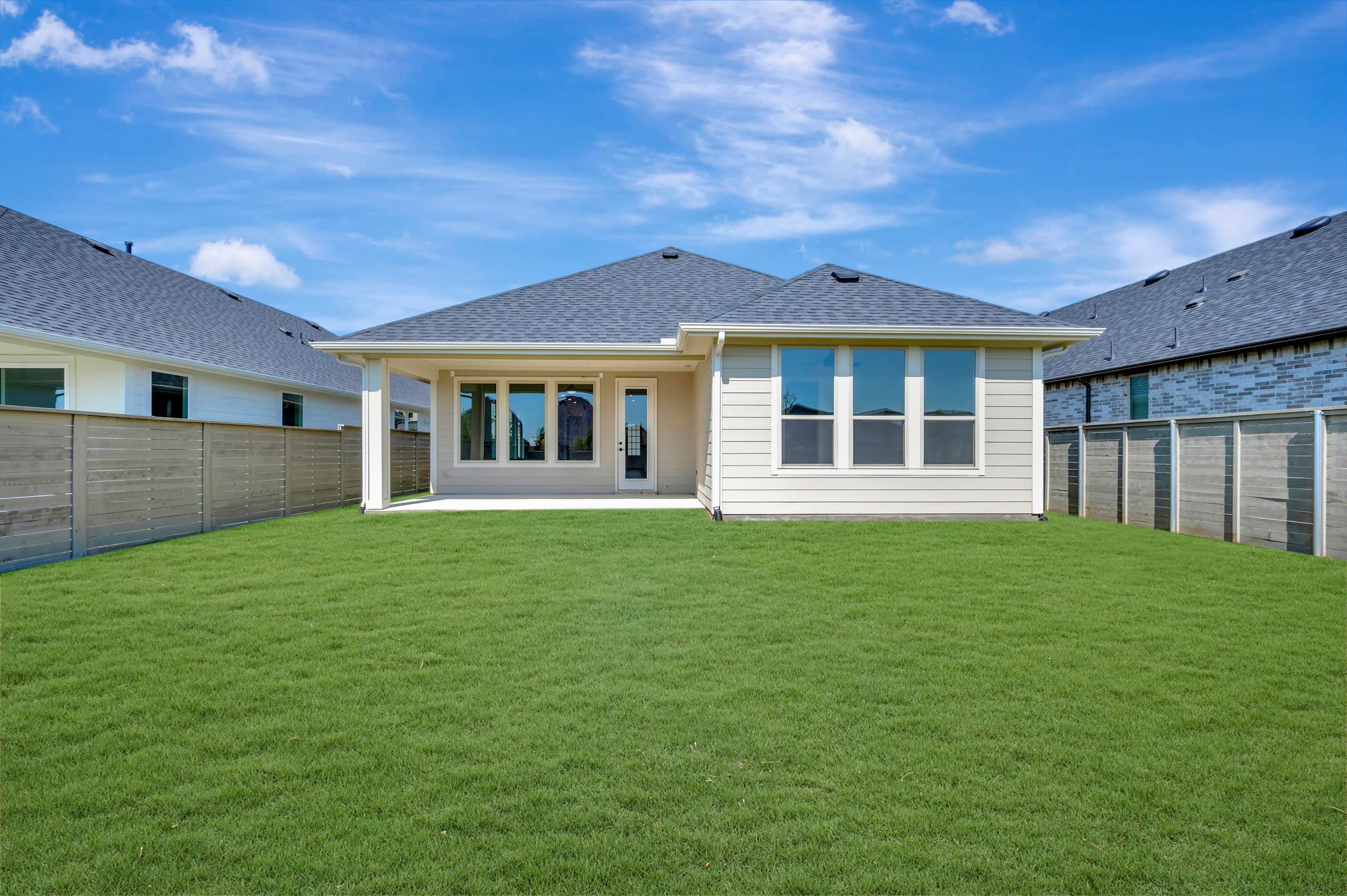 A freshly mowed green lawn stretches out before a beige house with large windows, while a light blue sky with scattered clouds forms the backdrop.