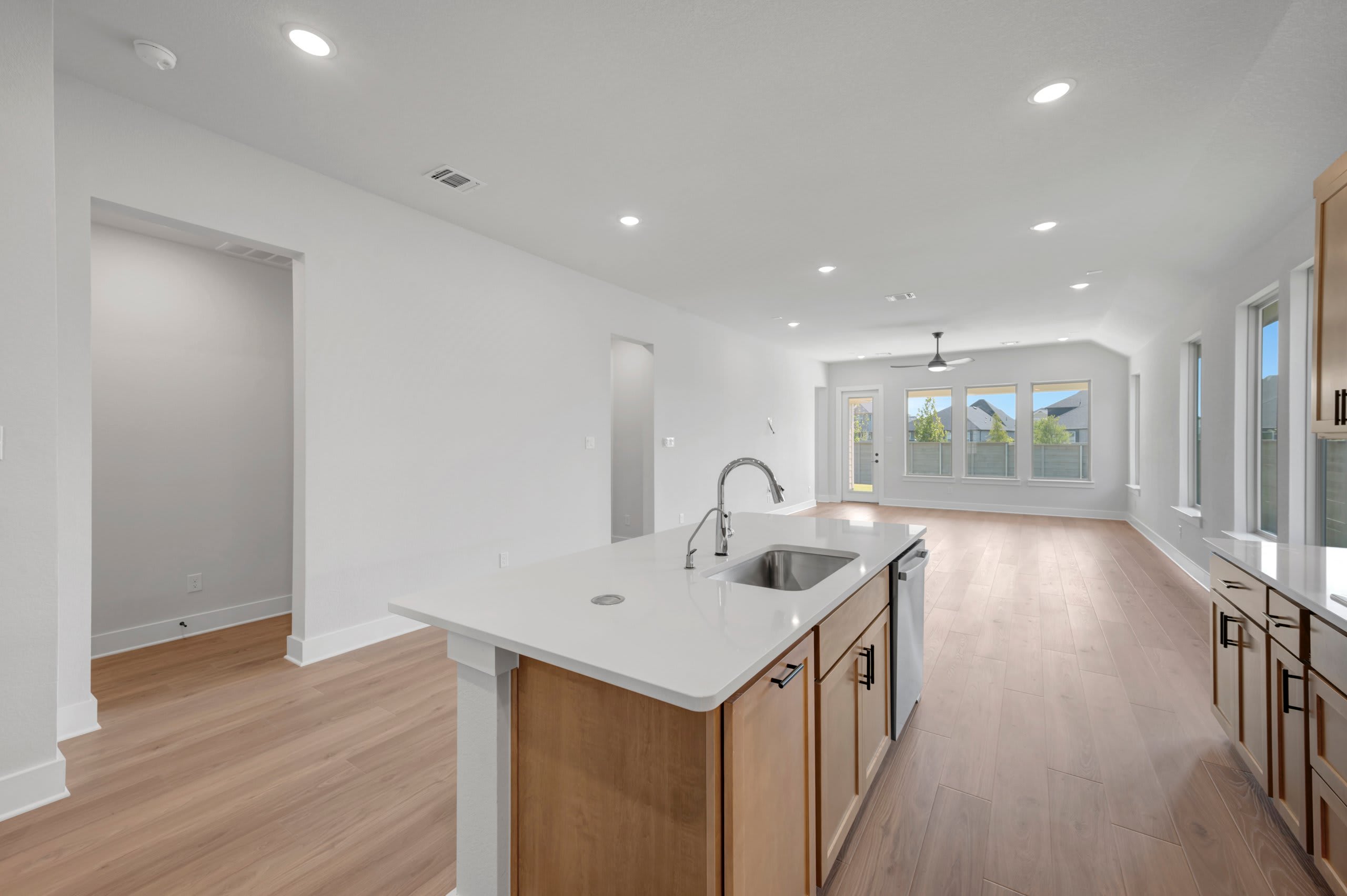 A modern kitchen island with a sink and faucet is in the foreground, while a spacious, open-plan living area with large windows and a ceiling fan is in the background.