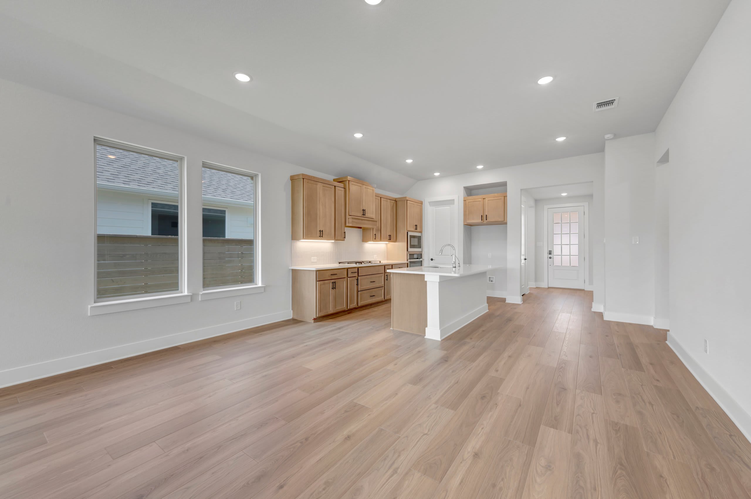 A spacious, modern living area with light wood flooring in the foreground opens to a bright kitchen with light wood cabinets and white countertops in the background.