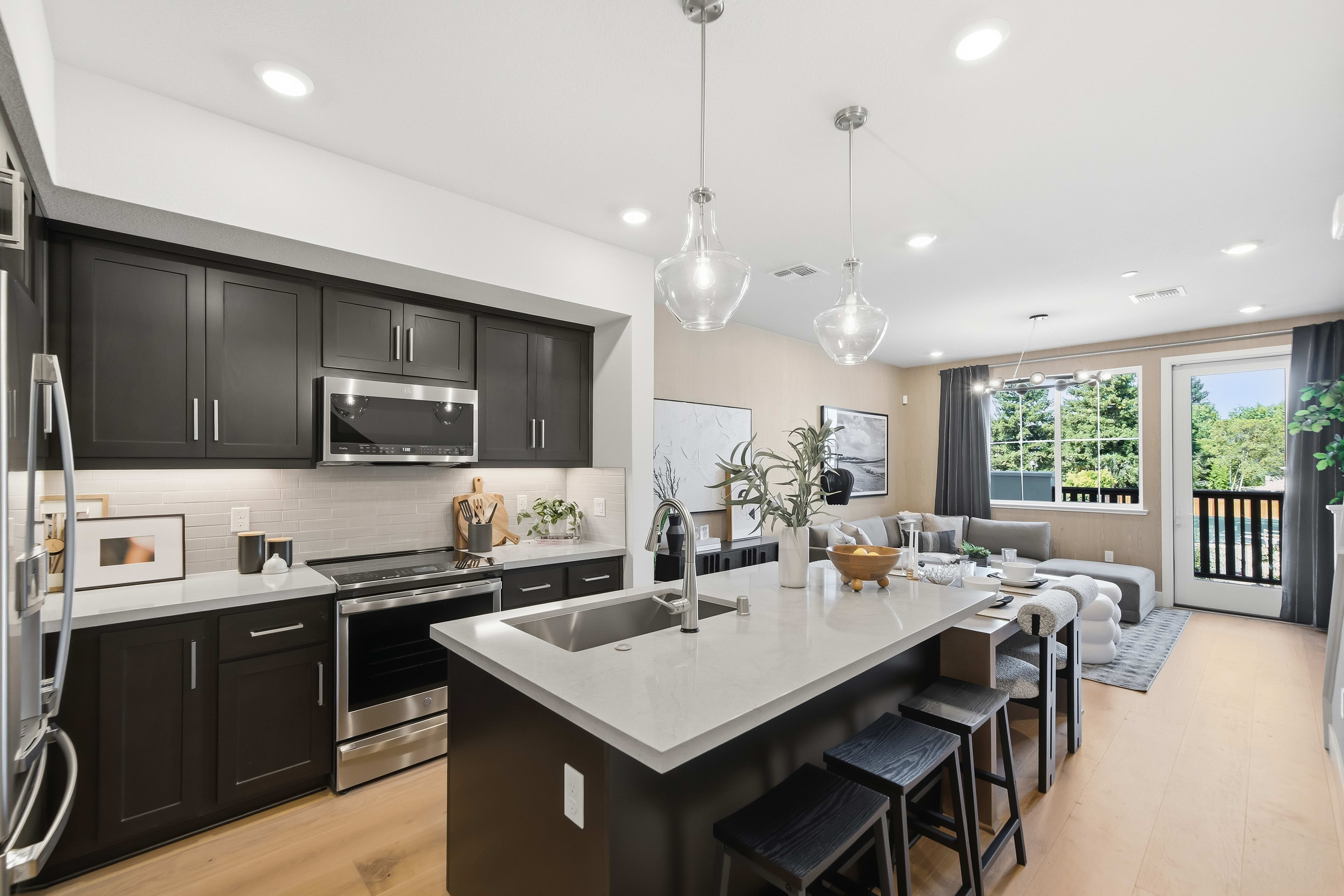 A modern kitchen with a large island in the foreground opens to a spacious living area with a sectional sofa and large windows in the background.