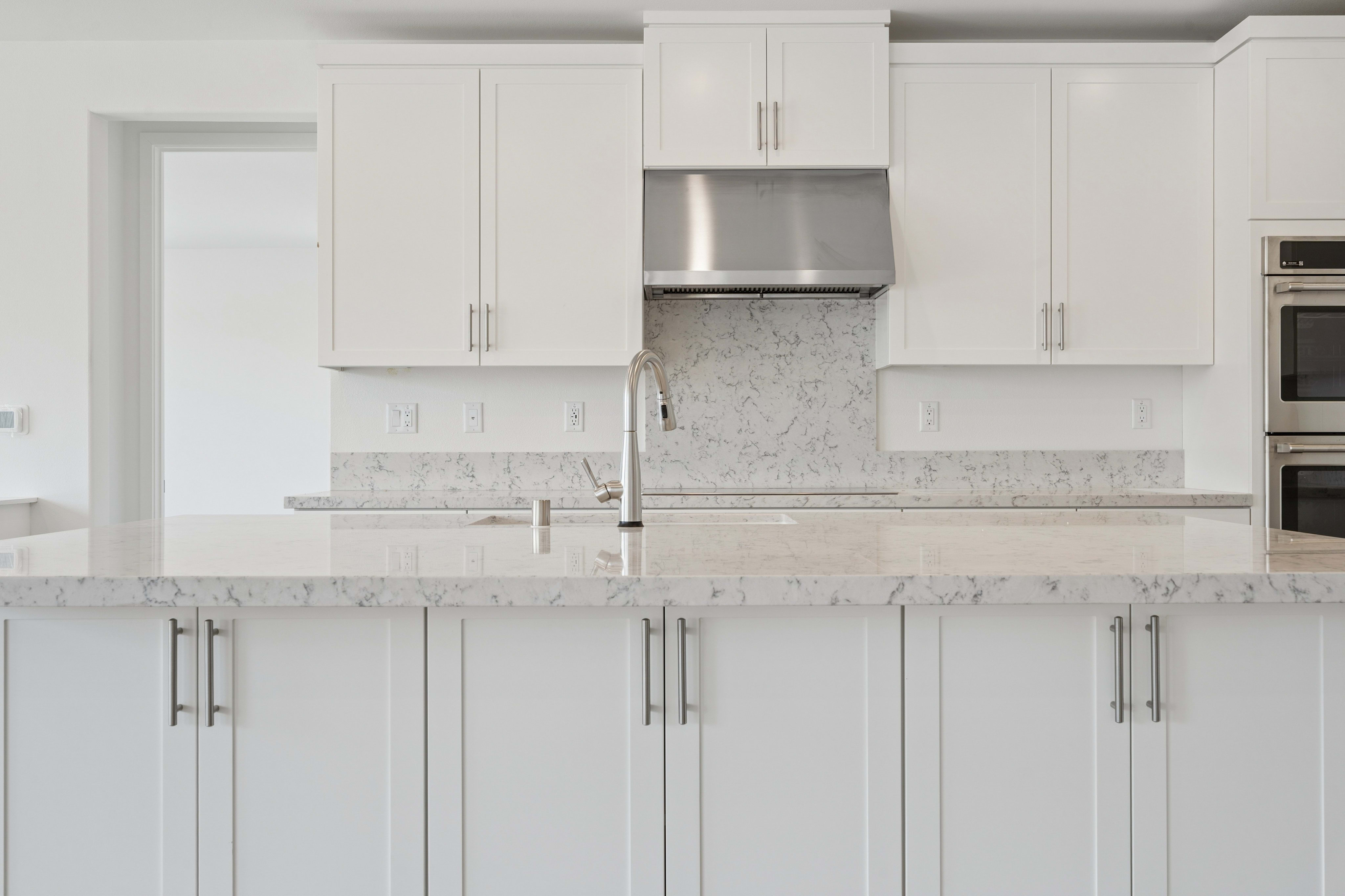 A modern white kitchen with a marble countertop in the foreground and white cabinets and stainless steel appliances in the background.
