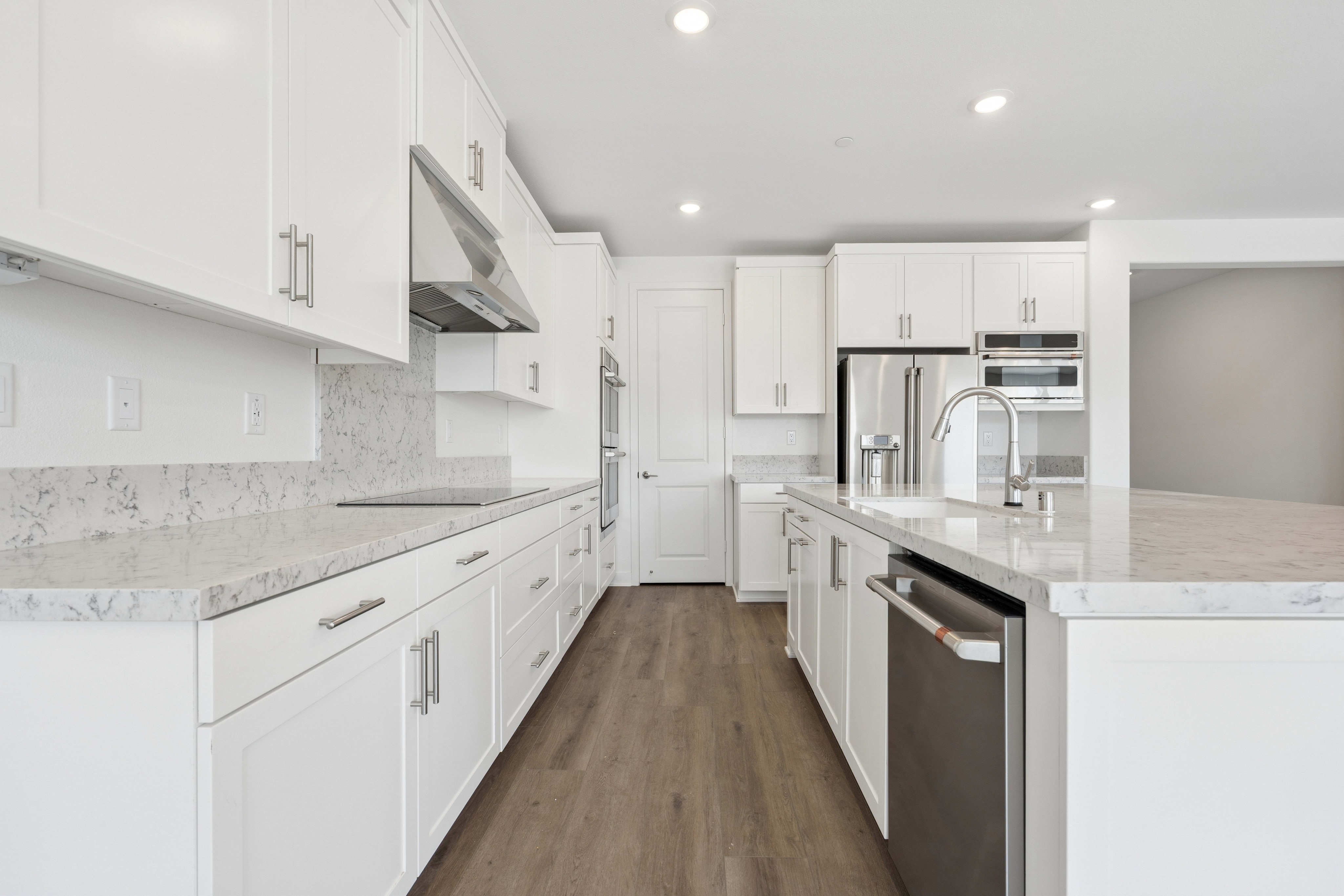A modern white kitchen with a granite countertop and stainless steel appliances is visible, with a doorway and a refrigerator in the background.