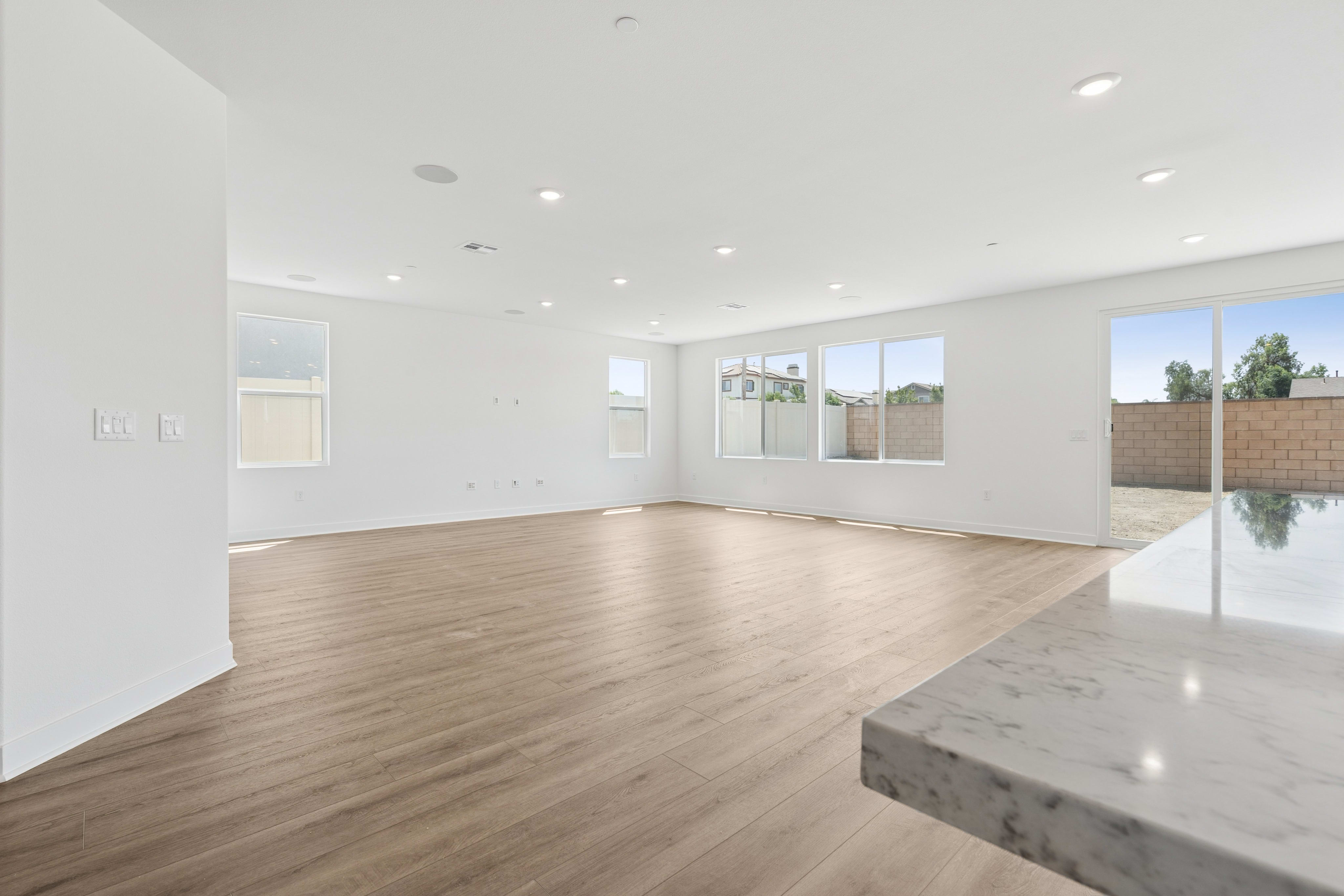 A bright, empty living room with light wood floors in the foreground and large windows looking out onto a backyard with trees and a brick wall in the background.