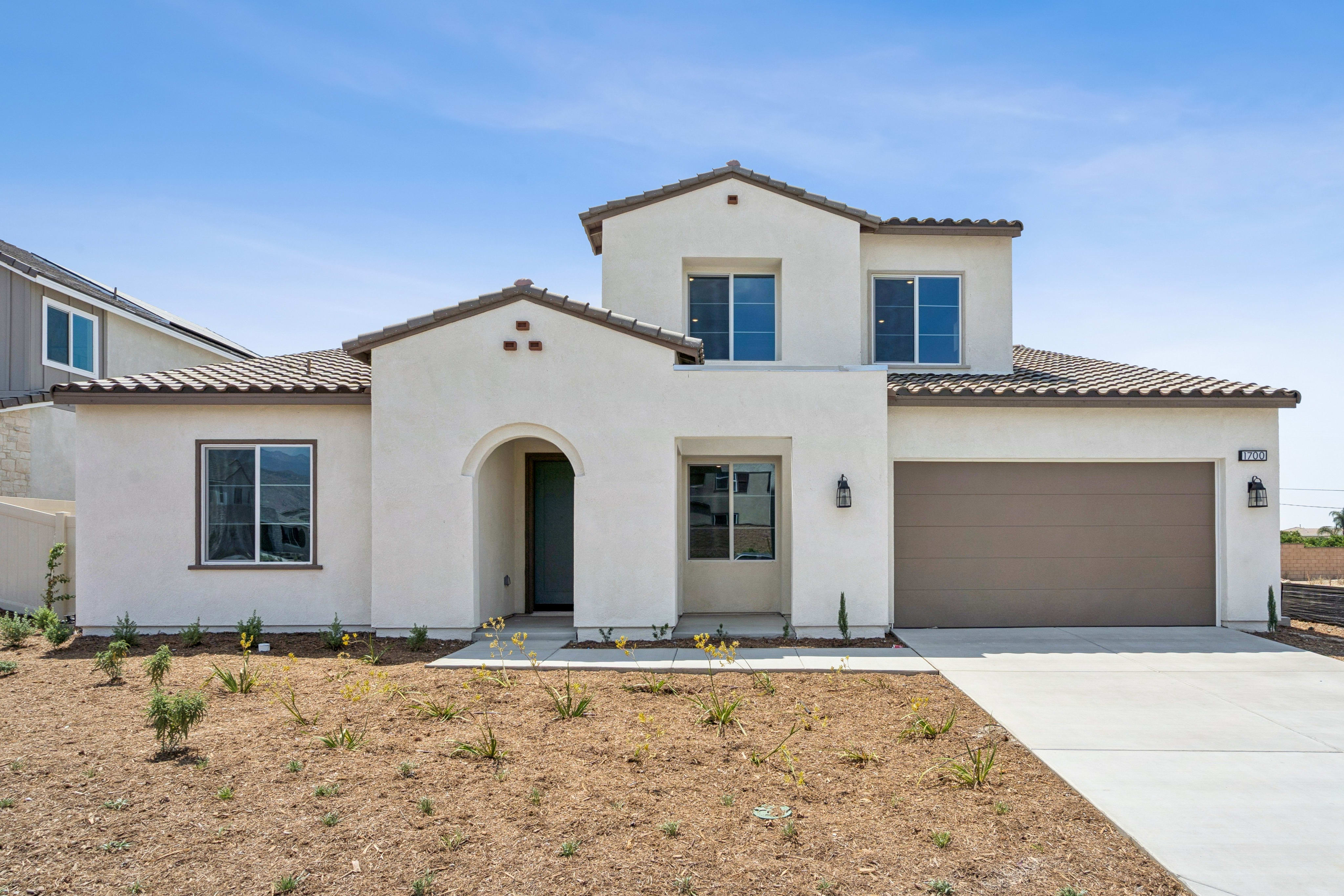 A modern stucco house with a light brown garage door sits on a dry, landscaped lawn under a clear blue sky.